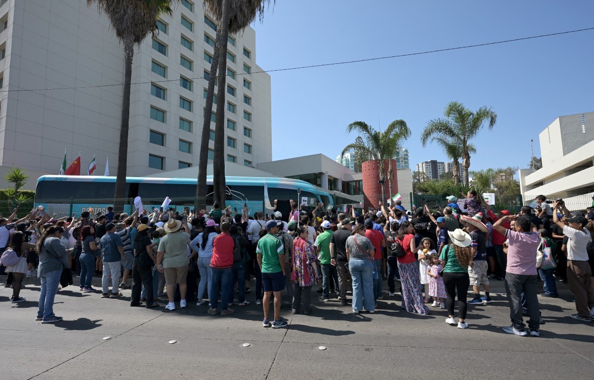 Multidão de pessoas, algumas com chapéus, reunidas em uma rua, com um ônibus azul e um prédio alto ao fundo, sob céu azul claro