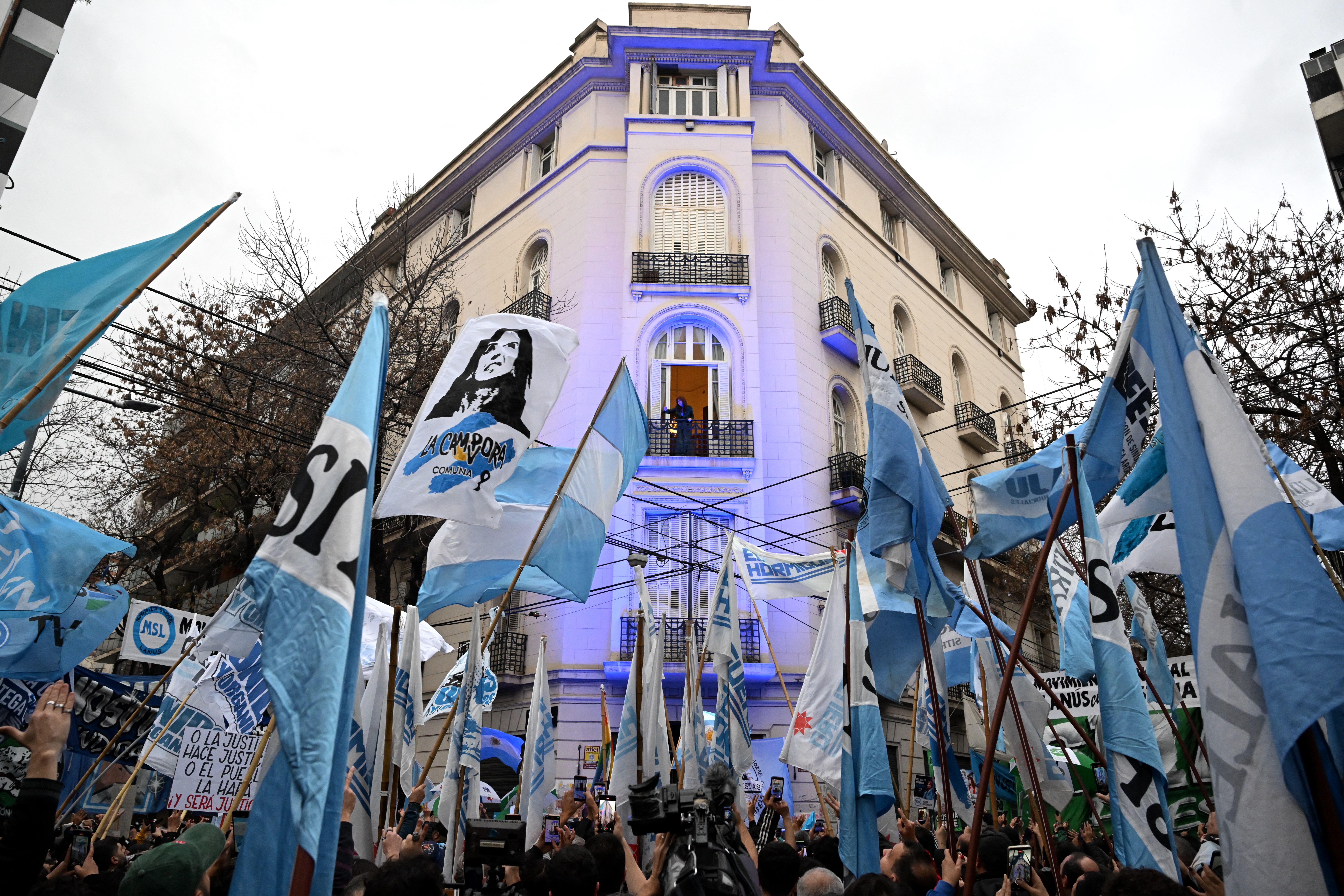 Multidão de pessoas com bandeiras azuis e brancas, algumas com o rosto de Eva Perón, em frente a um prédio branco iluminado por luzes azuis, com uma pessoa no balcão central. O céu está nublado A ex-presidente argentina (2007-2015) Cristina Kirchner aparece na varanda de sua casa enquanto permanece em prisão domiciliar em Buenos Aires, em meio a ato de apoiadores. 10/06/ 2026 -