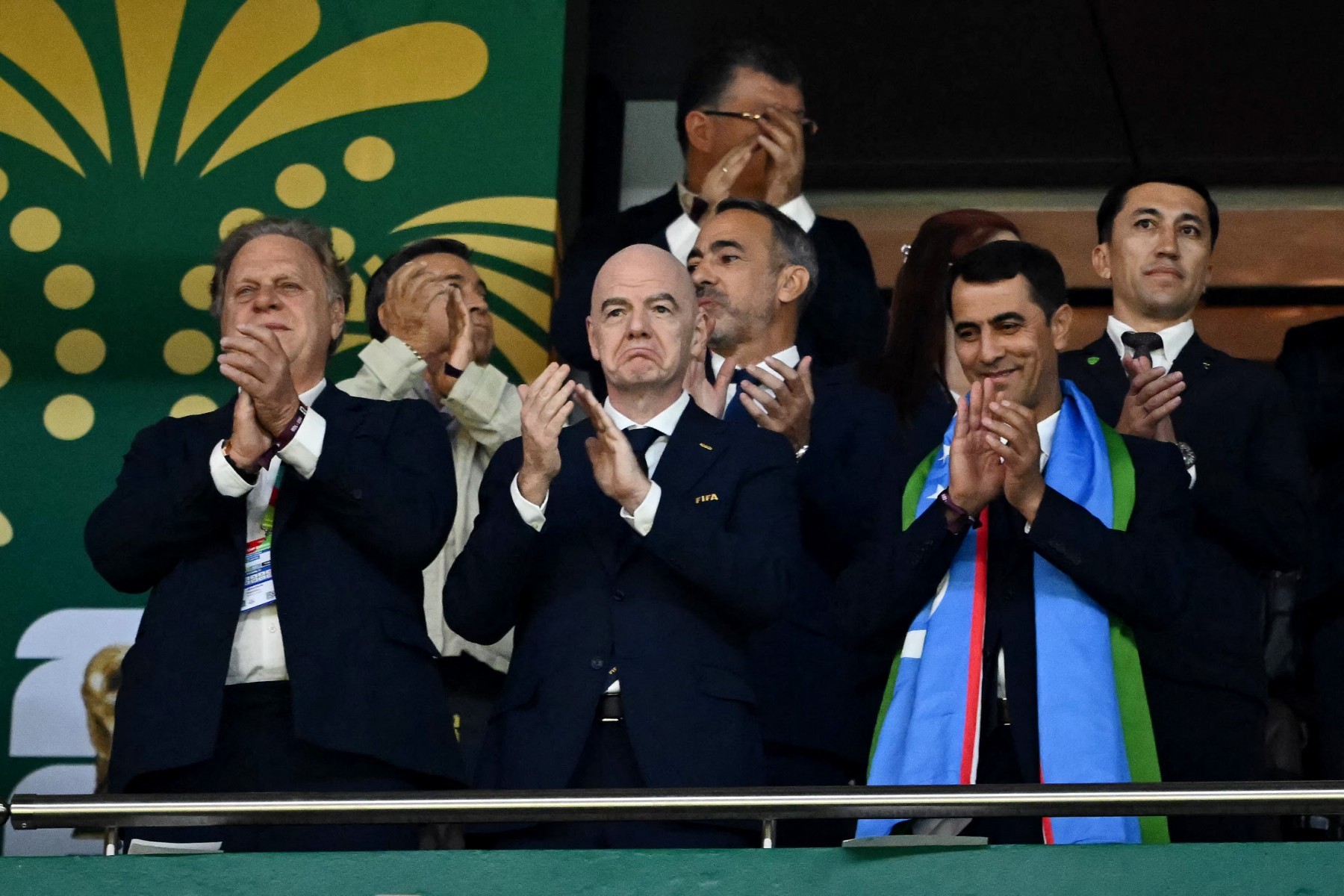 Homens de terno aplaudindo em uma arquibancada, com um fundo verde e amarelo. O presidente da FIFA, Gianni Infantino, está no centro, com expressão séria, enquanto outros sorriem. Um homem à direita usa um cachecol azul, verde e branco