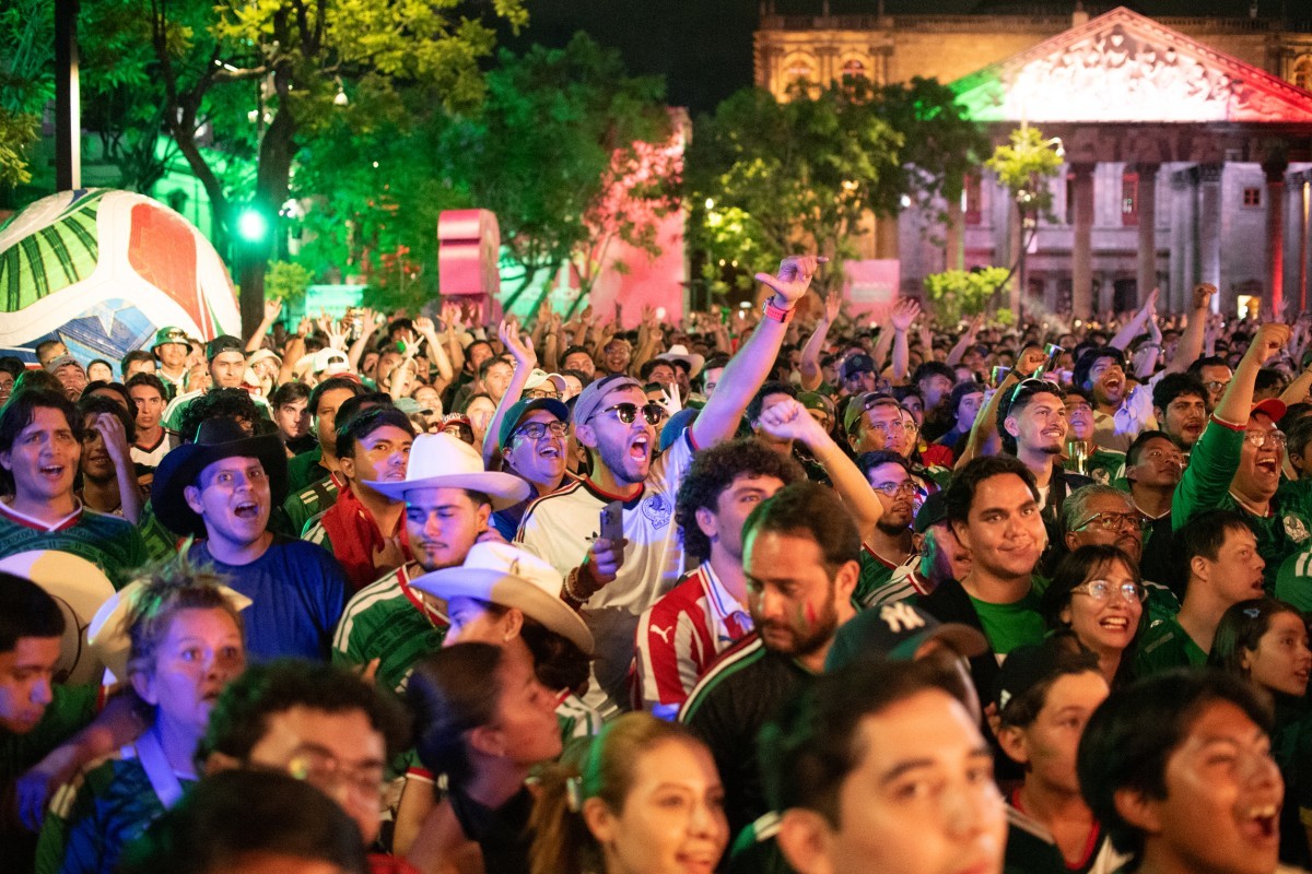 Multidão de pessoas animadas, algumas vestindo camisas de futebol e chapéus, comemorando à noite em uma praça iluminada por luzes verdes e vermelhas, cores da bandeira do México, projetadas em um edifício ao fundo