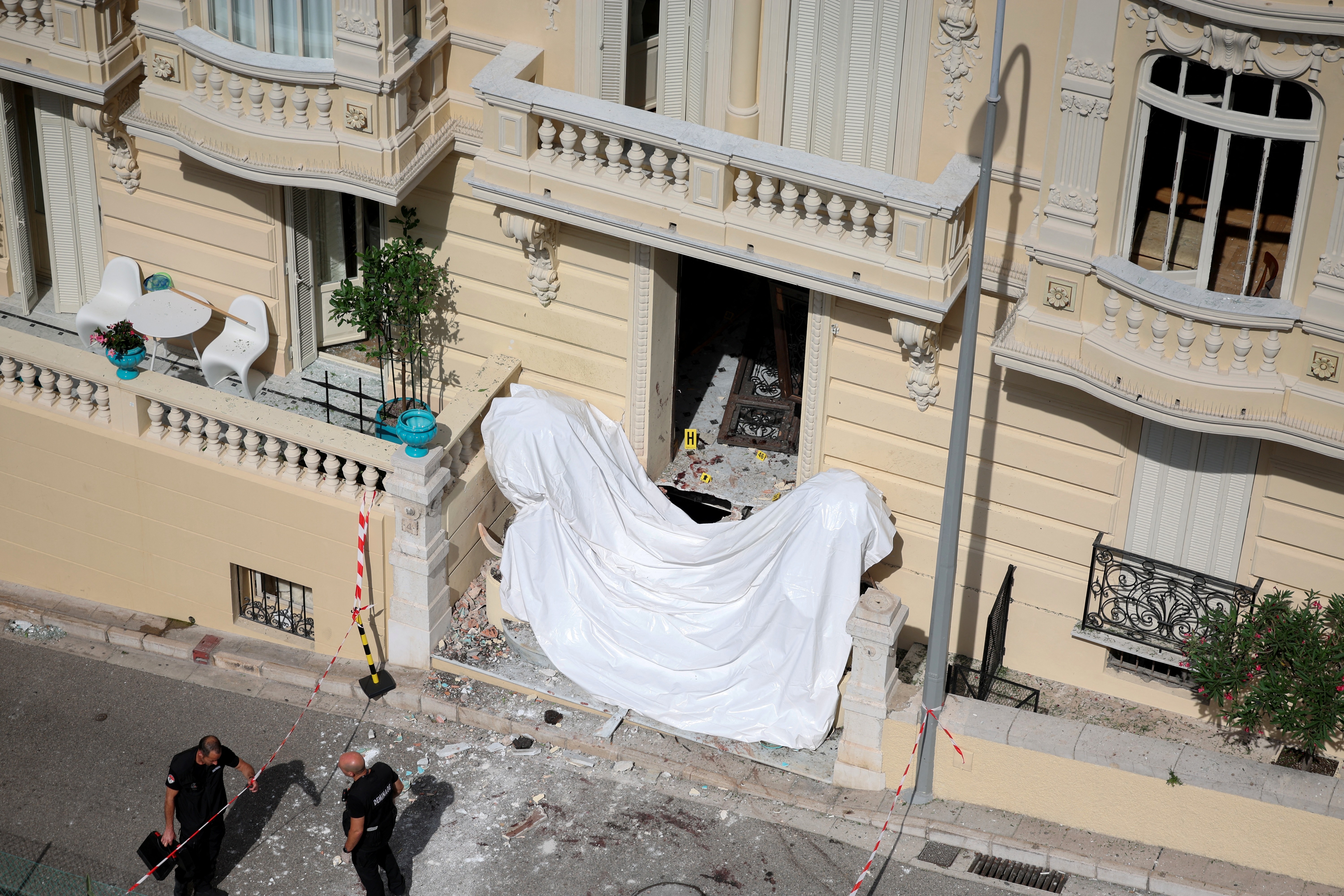 Fachada de prédio bege com varanda destruída, coberta por lona branca. Dois homens de uniforme escuro inspecionam a rua com detritos.