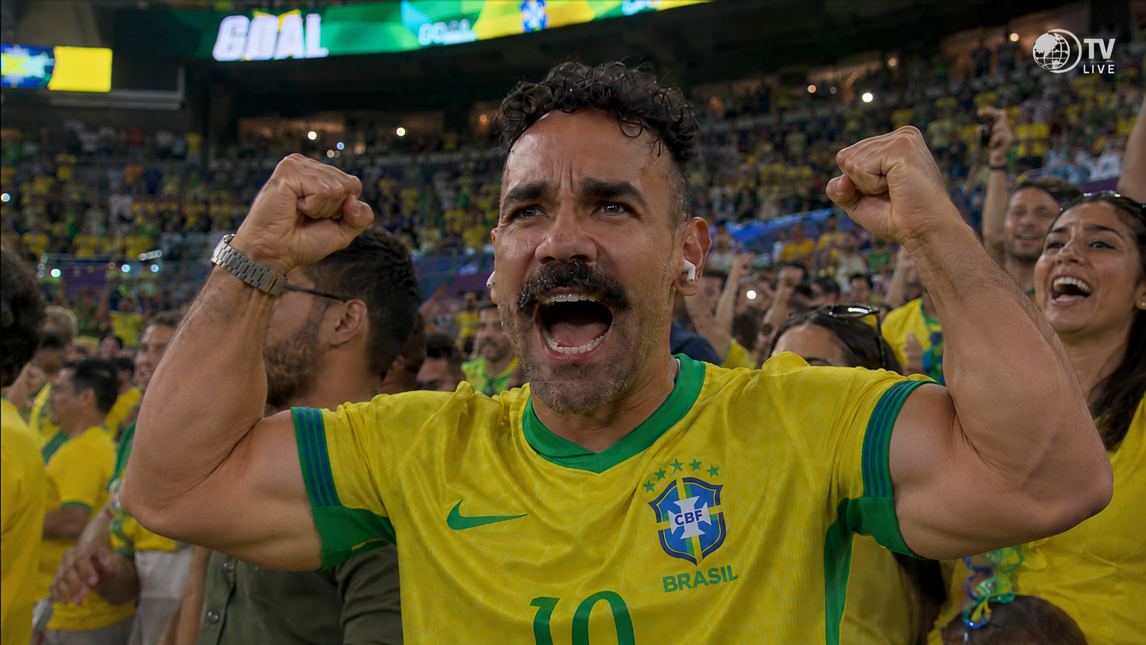 Homem de bigode e barba, vestindo camisa amarela da seleção brasileira, comemora um gol com os braços erguidos e punhos cerrados, boca aberta em grito de alegria. Ele está em um estádio lotado, com outros torcedores ao fundo, também celebrando. No placar, a palavra GOAL é visível