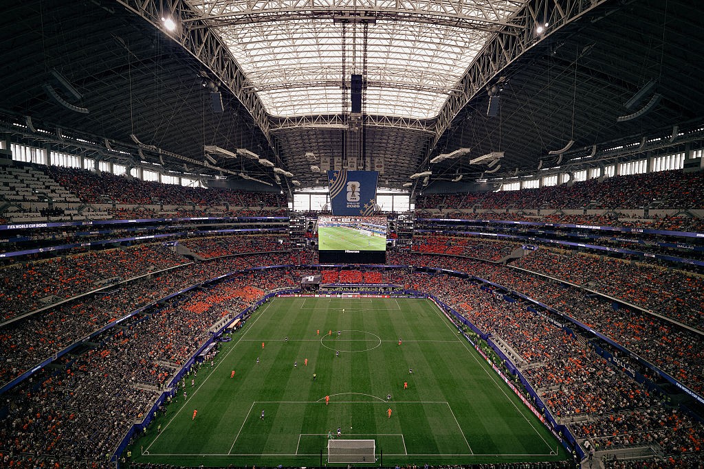 Vista aérea de um estádio de futebol lotado, com arquibancadas repletas de torcedores. O campo verde vibrante está no centro, com jogadores em campo. Um telão grande exibe a partida, e a estrutura metálica do teto é visível