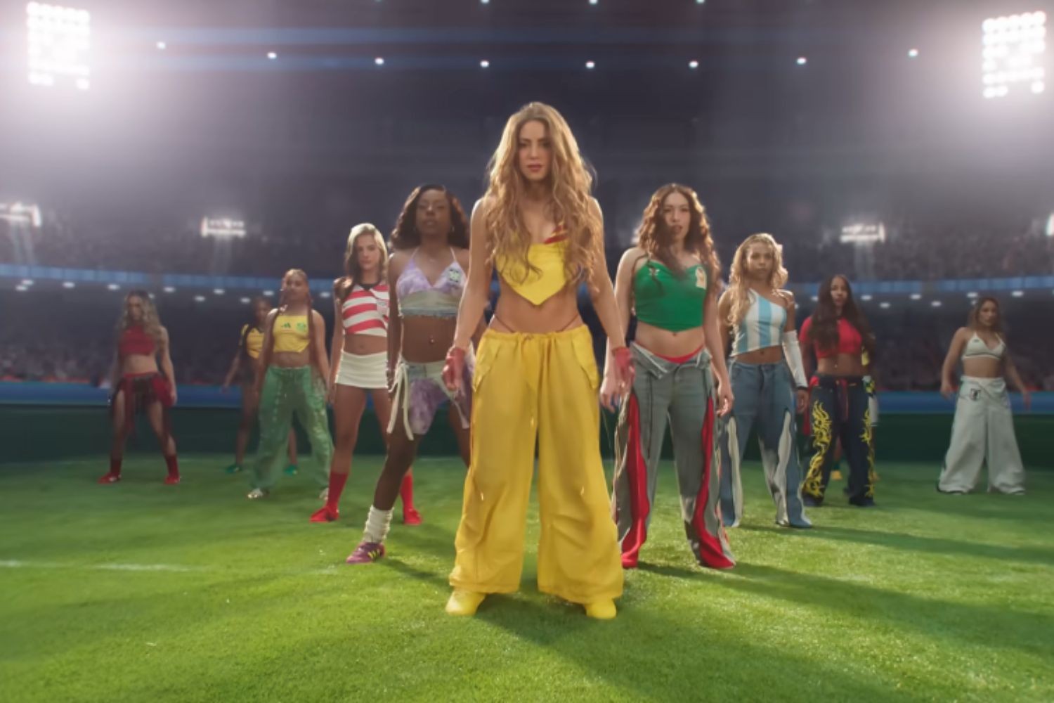 Shakira com top e calça amarela, cercada por mulheres em roupas coloridas, em um campo de futebol iluminado por holofotes