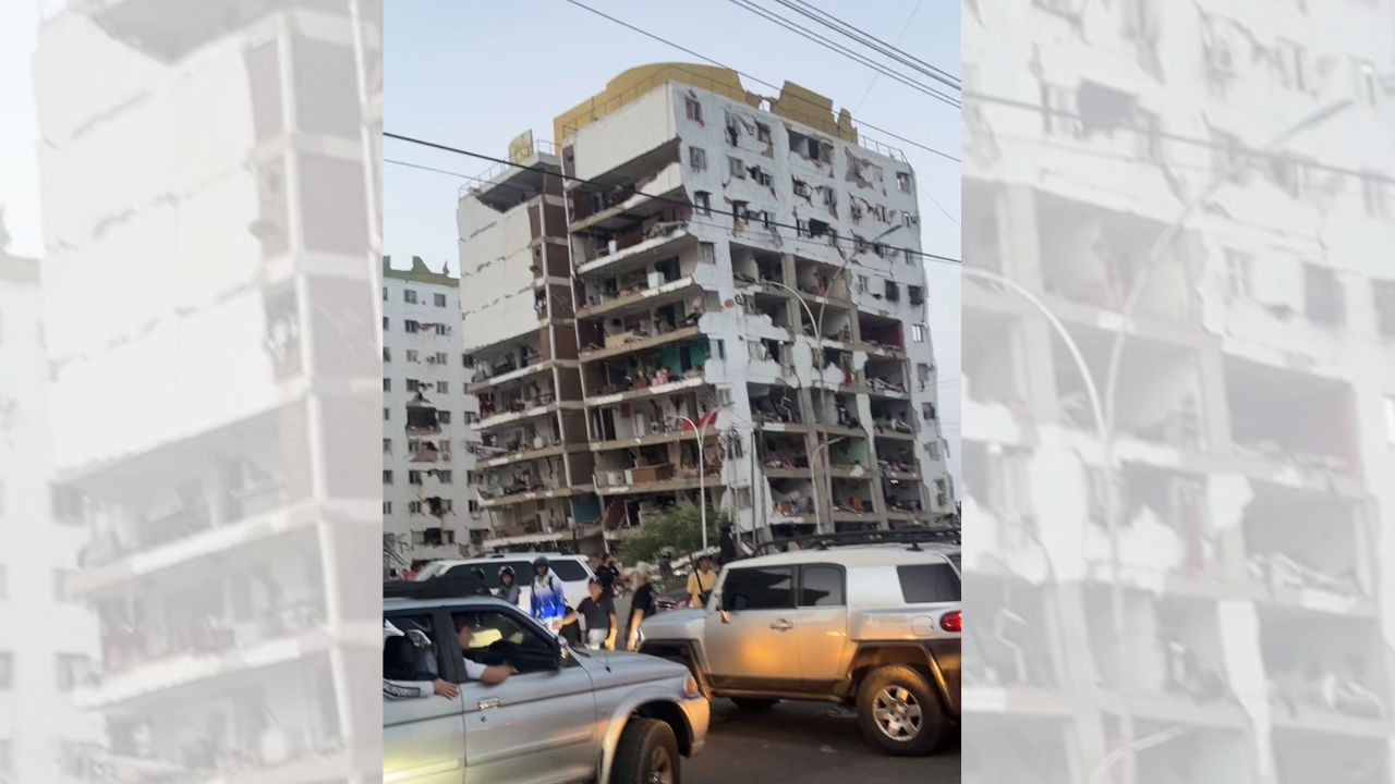Prédio de vários andares com fachada parcialmente destruída, revelando o interior dos apartamentos. Carros e pessoas transitam na rua em frente, sob um céu claro