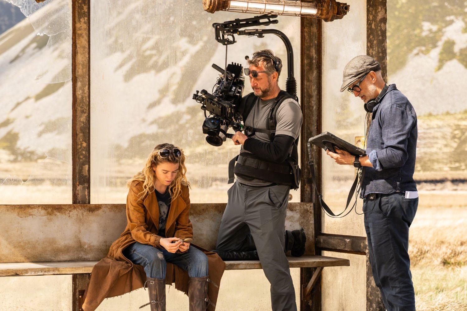 Três pessoas em um set de filmagem rústico. Uma mulher loira sentada em um banco de madeira, olhando para o celular. Um homem com óculos escuros e colete de câmera segura uma câmera profissional. Outro homem de boné e óculos lê um tablet.