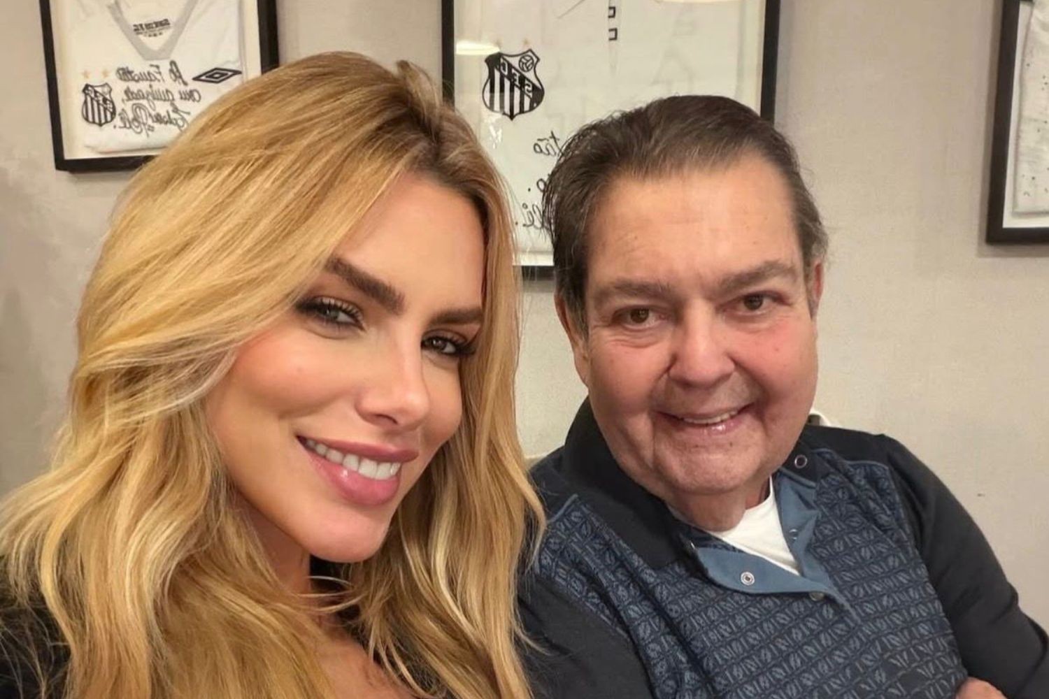 Apresentadora loira de cabelos longos e maquiagem leve sorri ao lado de um homem mais velho, de cabelos escuros e camisa azul estampada, ambos sorrindo para a c&acirc;mera. Ao fundo, camisas de futebol emolduradas.