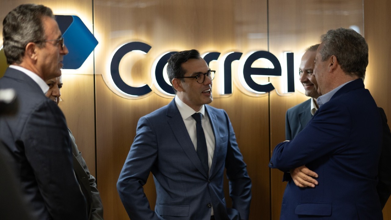 Quatro homens em ternos conversam em um ambiente corporativo. Ao centro, um homem de óculos e terno azul escuro sorri, olhando para a direita. Ao fundo, um letreiro luminoso com o nome Correios em azul e branco. À esquerda, um homem de perfil e outro parcialmente visível. À direita, dois homens de costas, um deles sorrindo.