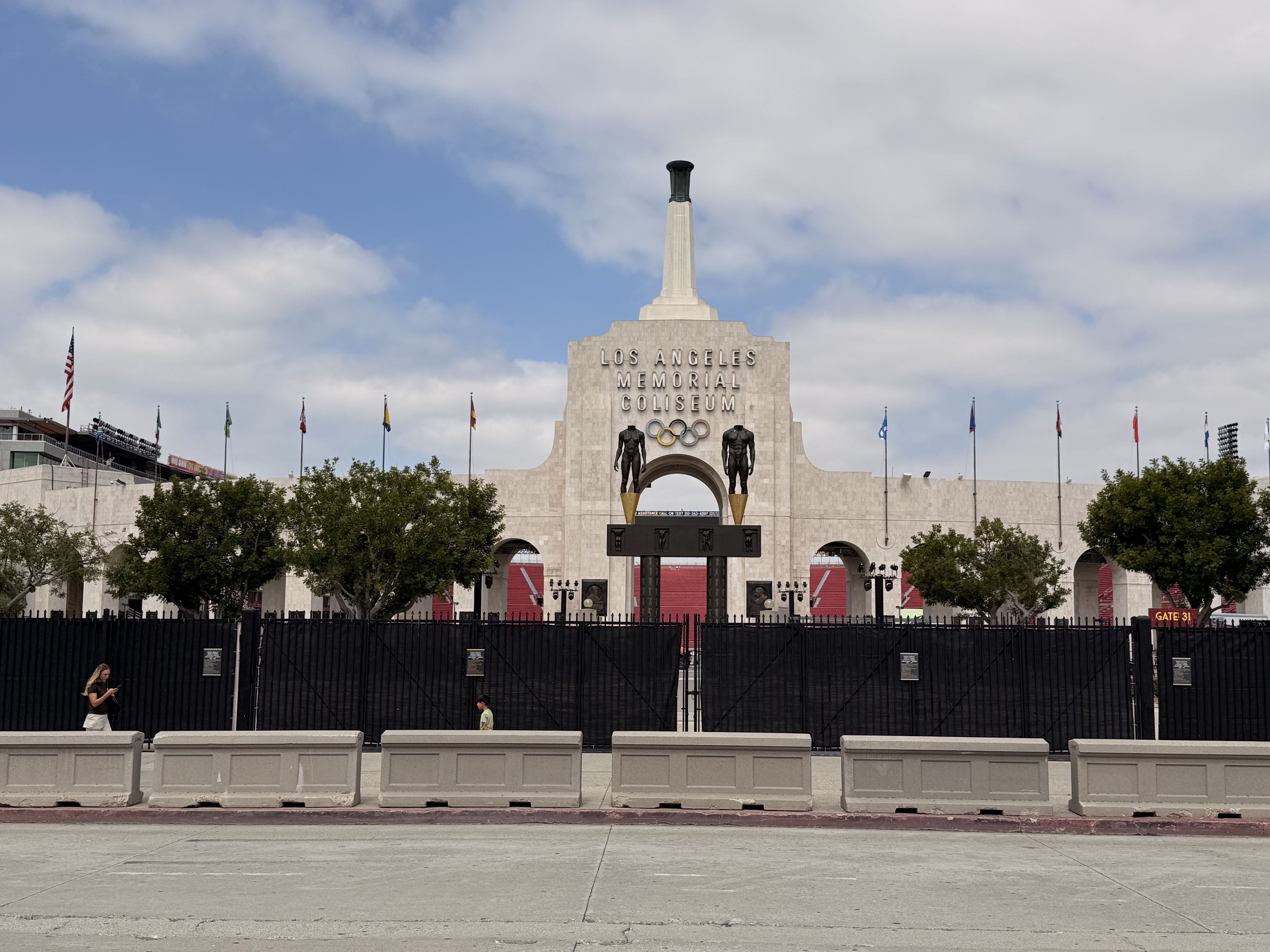 Fachada do Los Angeles Memorial Coliseum, com a torre central e o nome do estádio. Duas estátuas de atletas e os anéis olímpicos adornam a entrada. Uma cerca preta e barreiras de concreto cinzas separam o estádio da rua. O céu está parcialmente nublado, com algumas pessoas caminhando ao longe