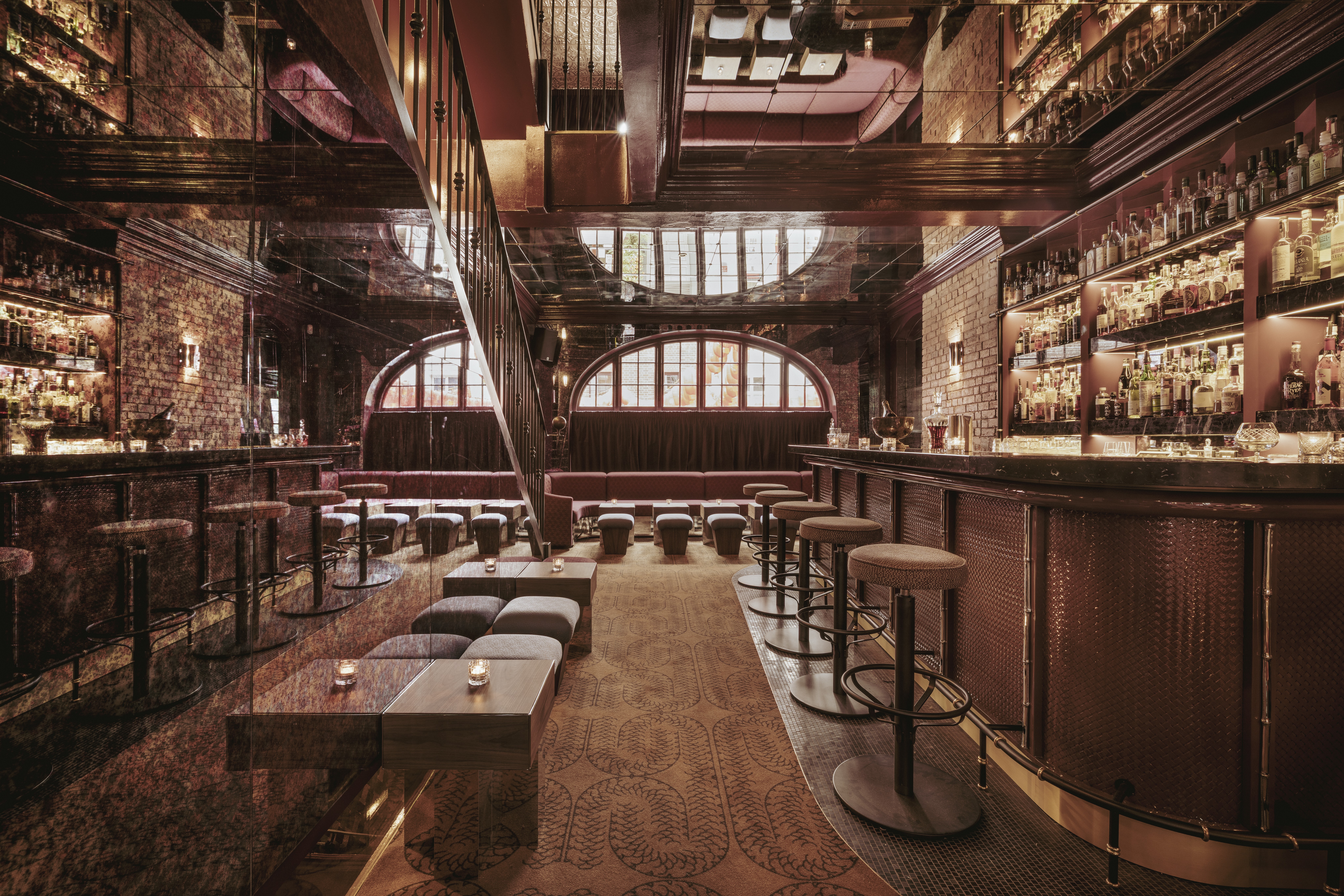 Interior de um bar sofisticado com iluminação baixa e tons terrosos. À direita, um balcão longo com banquetas estofadas e prateleiras cheias de garrafas. Ao fundo, janelas arqueadas e sofás vermelhos. Uma escada escura sobe à esquerda, e mesas baixas com velas estão no centro, sobre um tapete estampado. O teto espelhado reflete o ambiente, criando uma atmosfera aconchegante e elegante