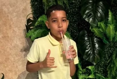 Menino de pele clara, cabelo curto, vestindo camisa polo amarela, bebendo um milkshake rosa com canudo. Ele faz um sinal de "hang loose" com a mão esquerda, em frente a uma parede decorada com folhagens verdes e uma parede marrom