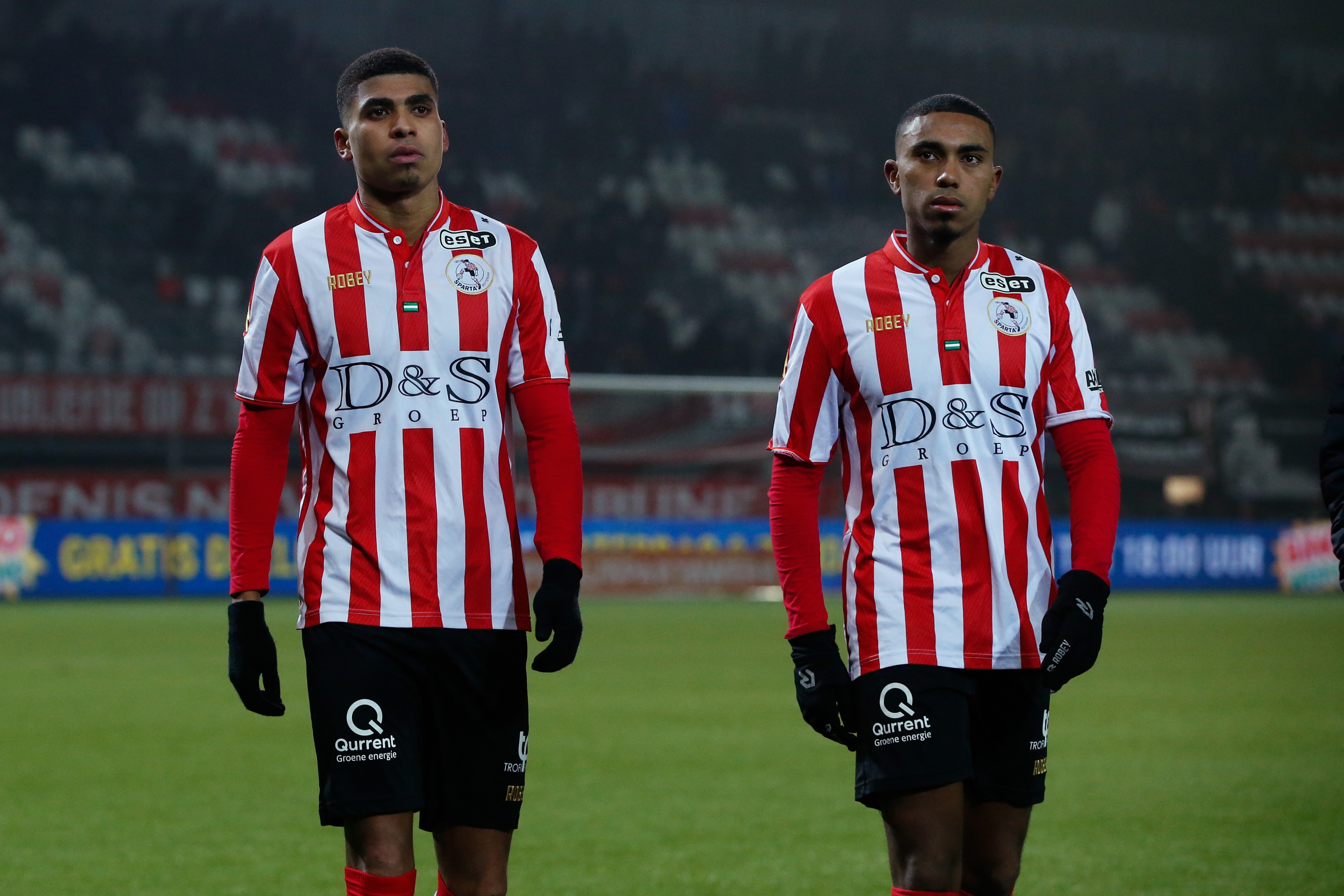 Dois jogadores de futebol, um à esquerda e outro à direita, vestem camisas listradas vermelhas e brancas, calções pretos e luvas pretas, em um campo de grama verde com arquibancadas ao fundo
