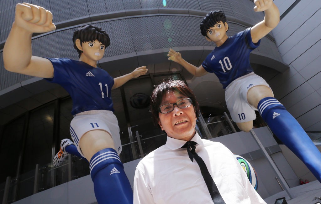 Um homem japonês de óculos e camisa branca com gravata preta sorri para a câmera, ladeado por duas estátuas de jogadores de futebol de anime em uniformes azuis e brancos, com os números 10 e 11, em pose de chute