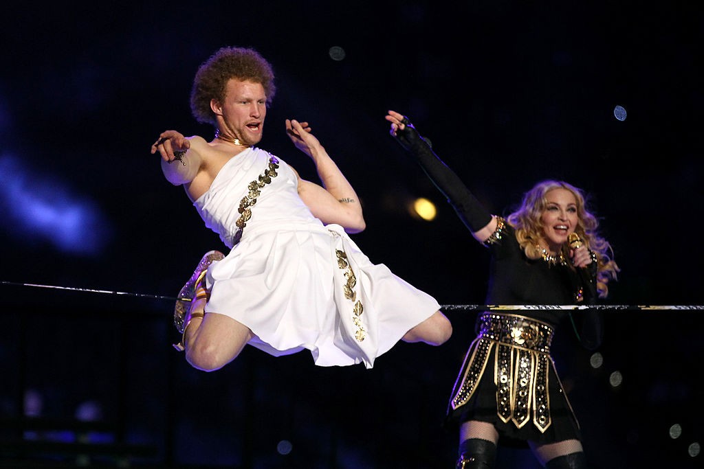 Um homem com cabelo afro e túnica branca com detalhes dourados, suspenso no ar, e Madonna com cabelo loiro, blusa preta e saia dourada, cantando em um microfone dourado, em um palco escuro com luzes azuis INDIANAPOLIS, IN - FEBRUARY 05: Madonna (R) performs during the Bridgestone Super Bowl XLVI Halftime Show at Lucas Oil Stadium on February 5, 2012 in Indianapolis, Indiana. (Photo by Al Bello/Getty Images)