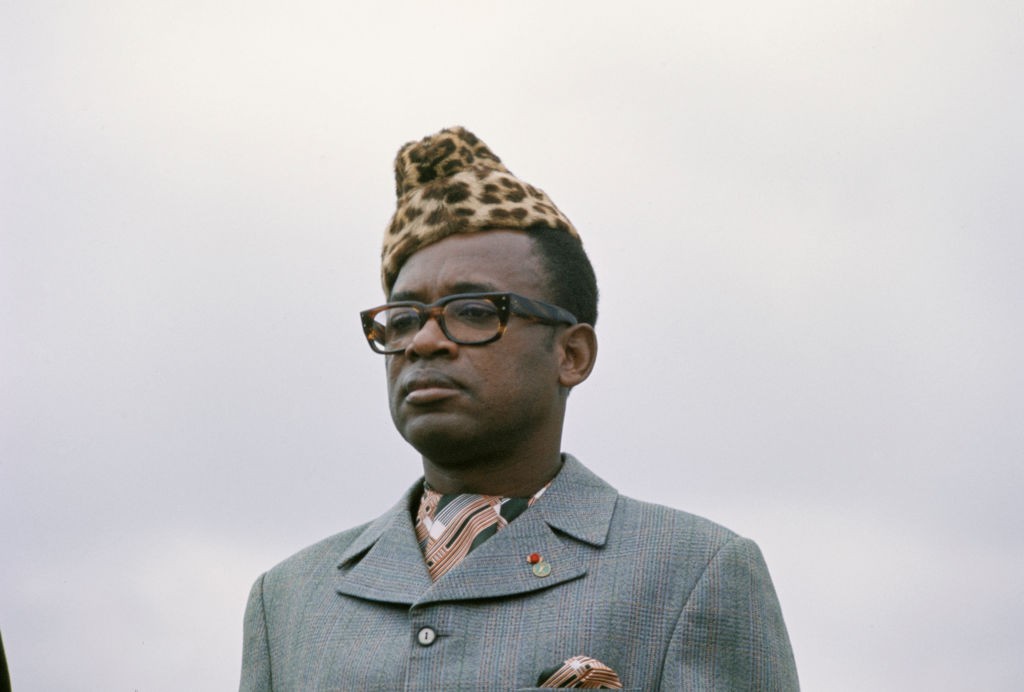 Homem negro de óculos e chapéu de pele de leopardo, vestindo um terno cinza e lenço estampado, com expressão séria, olhando para a frente