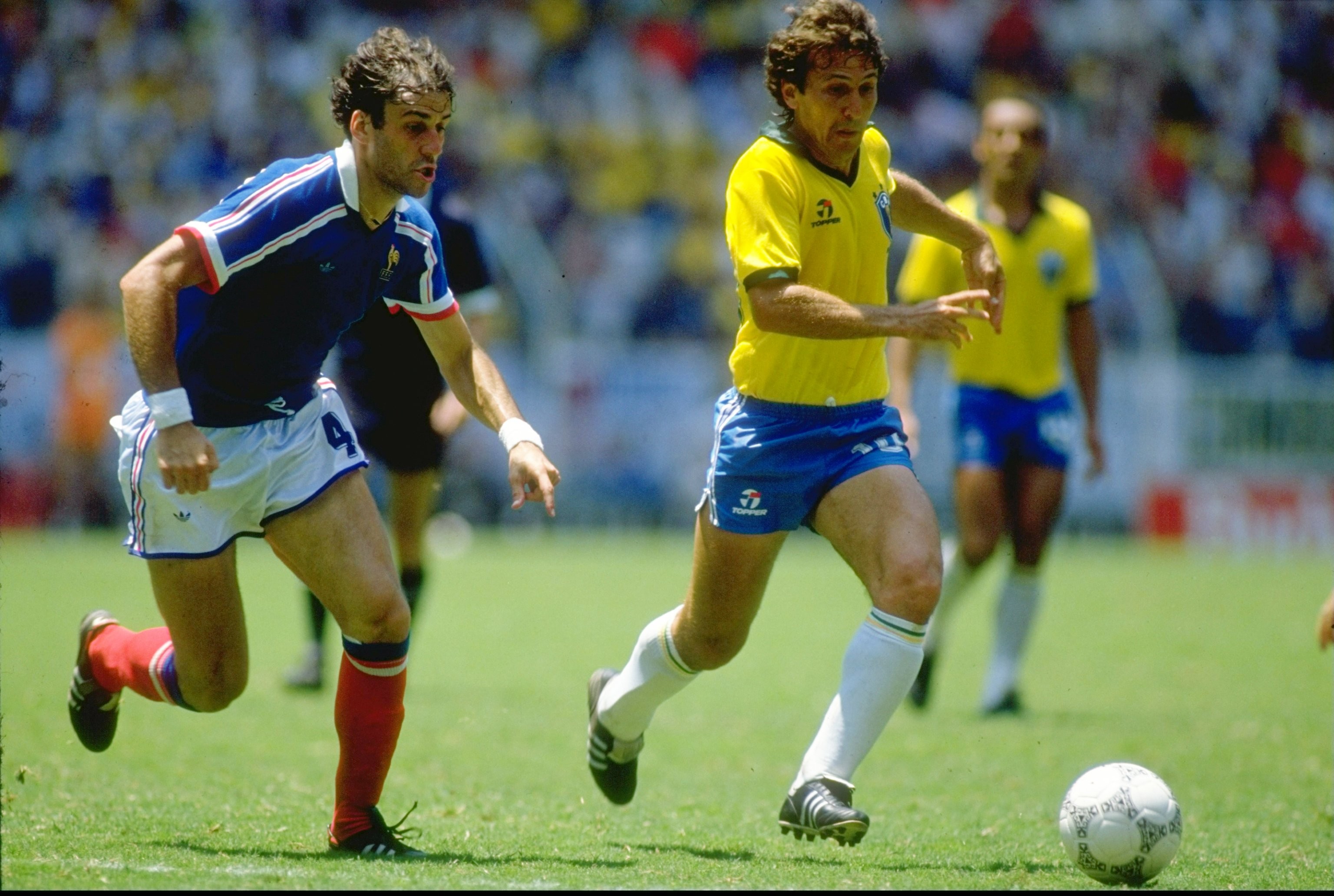 Dois jogadores de futebol correm em campo verde. Um, à esquerda, veste camisa azul, shorts brancos e meias vermelhas. O outro, à direita, usa camisa amarela, shorts azuis e meias brancas, com uma bola de futebol branca aos seus pés. Outros jogadores e torcedores desfocados ao fundo