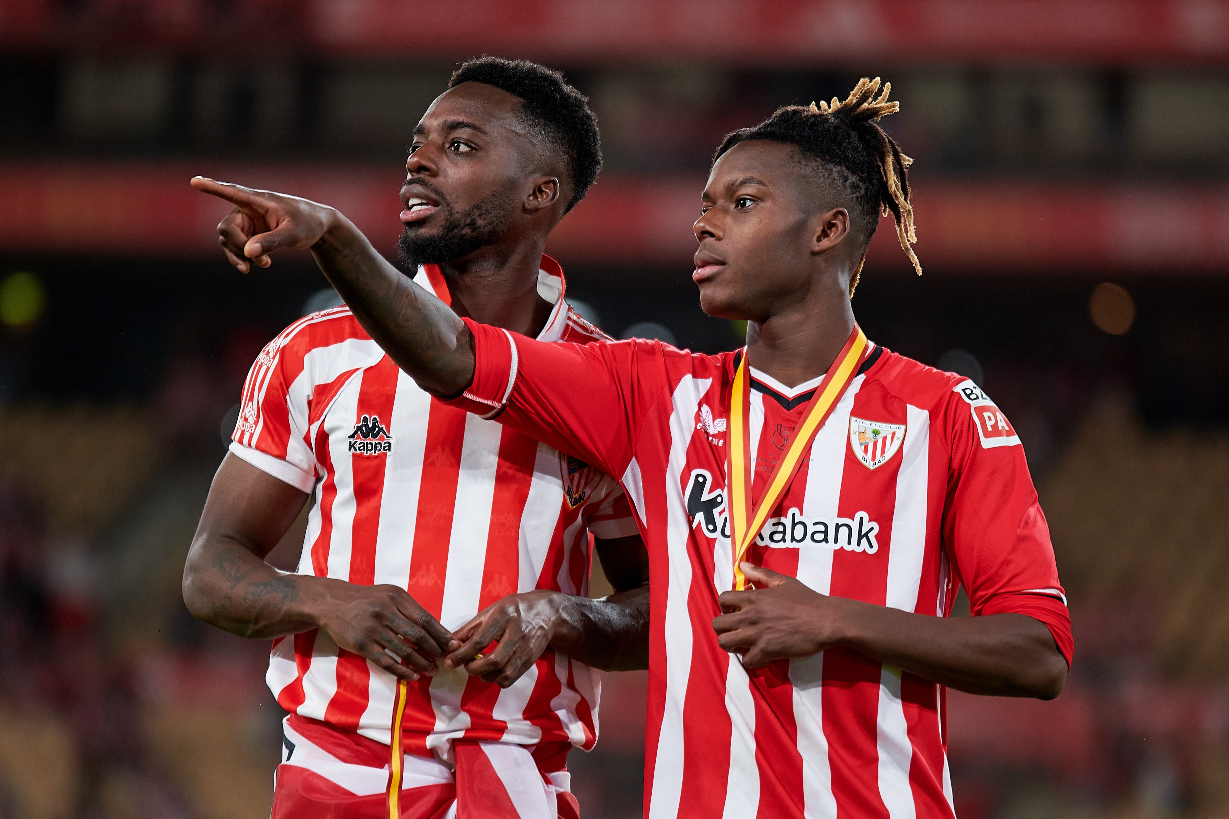Dois jogadores de futebol, um apontando para a direita e outro com uma medalha de ouro no pescoço, ambos vestindo camisas listradas vermelhas e brancas