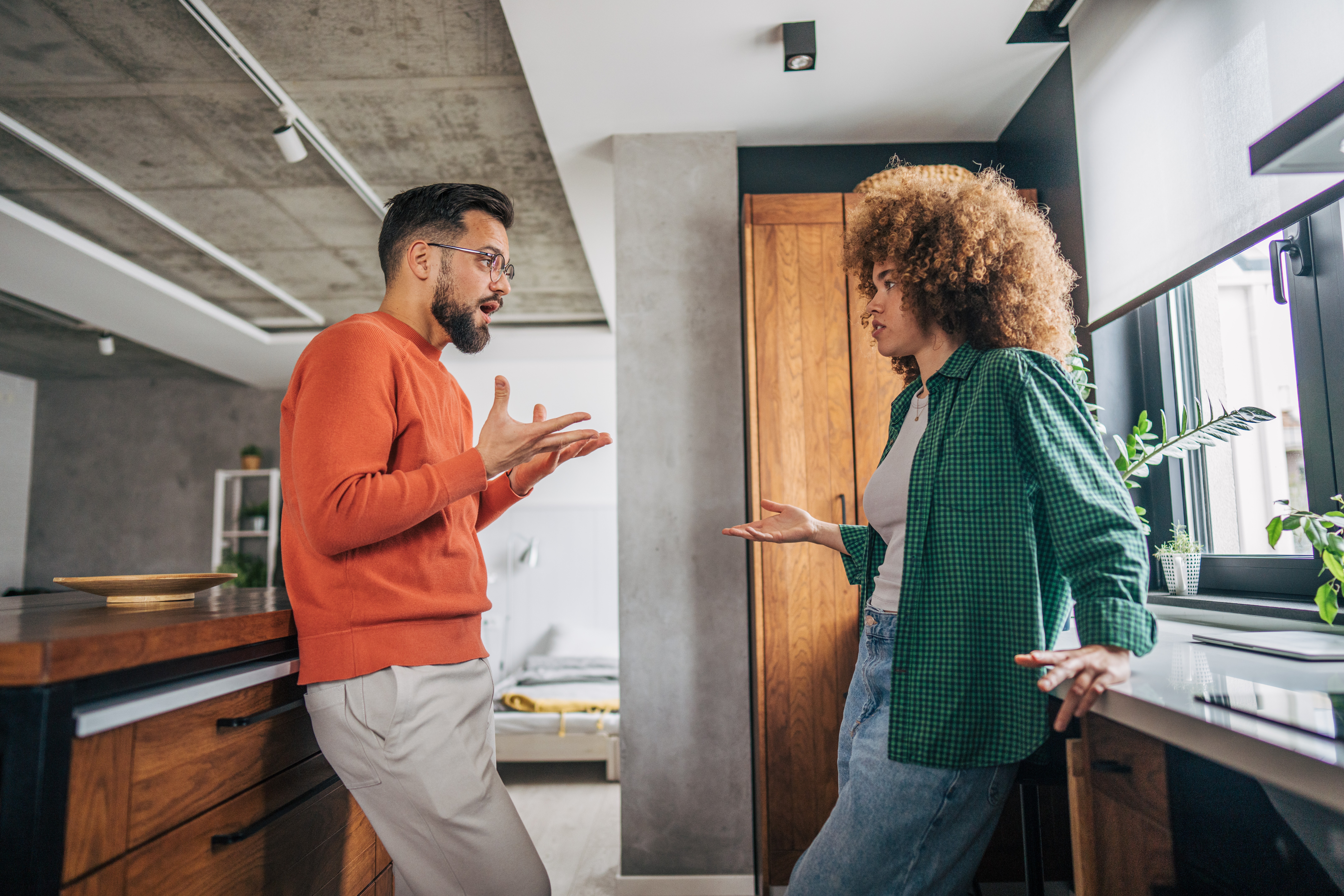 Um homem e uma mulher, ambos com expressões sérias e gesticulando com as mãos abertas, discutem em uma cozinha moderna. Ele veste suéter laranja e calça clara, ela usa camisa xadrez verde e jeans.