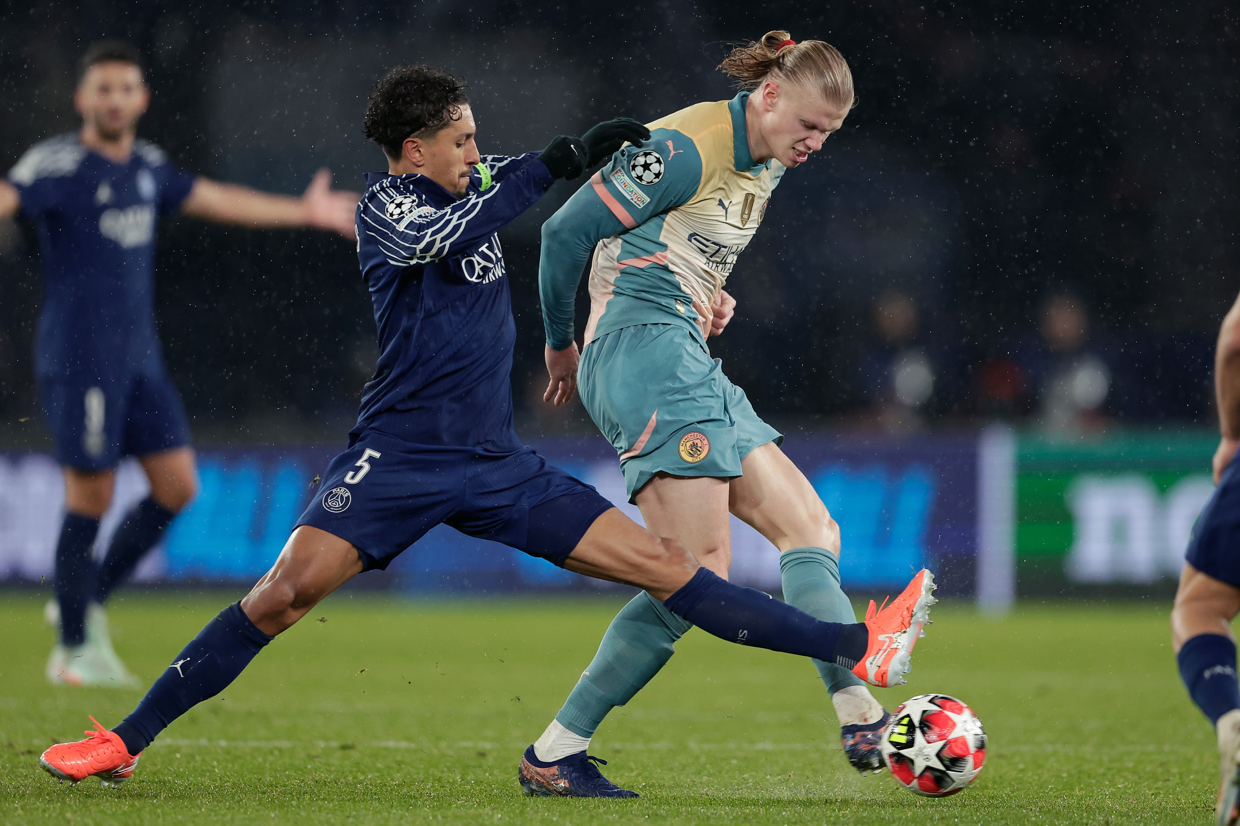 Dois jogadores de futebol em campo. Um, de camisa azul escura com número 5, tenta desarmar o outro, de camisa verde-água e cabelo loiro preso, que domina a bola vermelha e branca com o pé direito. Outro jogador azul desfocado ao fundo.