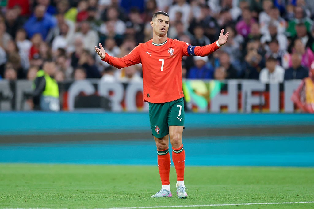 Cristiano Ronaldo, jogador de futebol, em campo com uniforme vermelho e verde, número 7, braços abertos em gesto de questionamento, em frente a uma plateia desfocada
