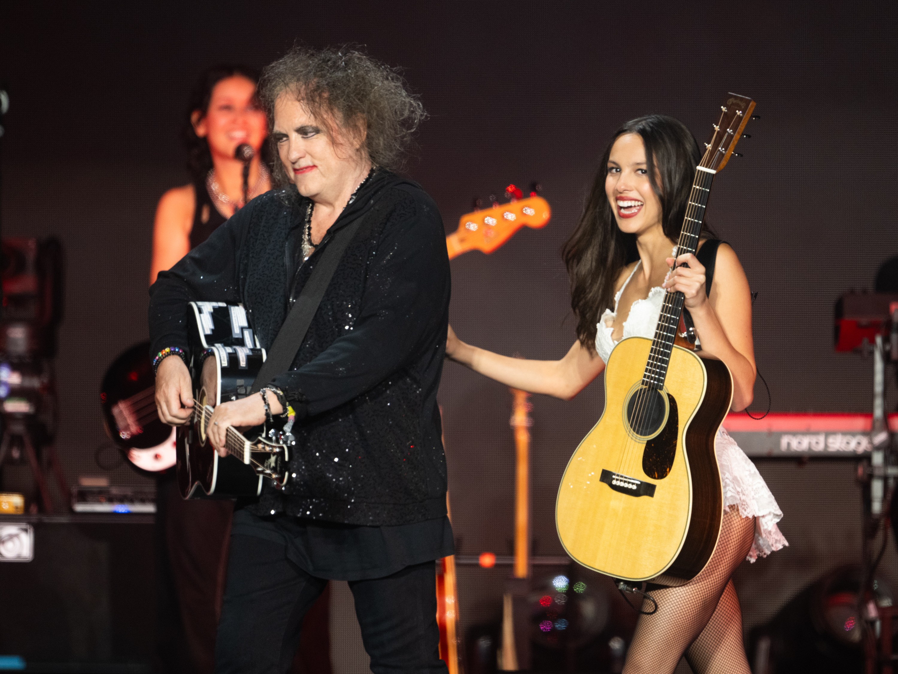Robert Smith, com cabelo volumoso e maquiagem escura, toca violão preto ao lado de uma mulher sorridente, de vestido branco e meias arrastão, segurando um violão claro. Outra mulher canta ao fundo, em um palco escuro com instrumentos musicais