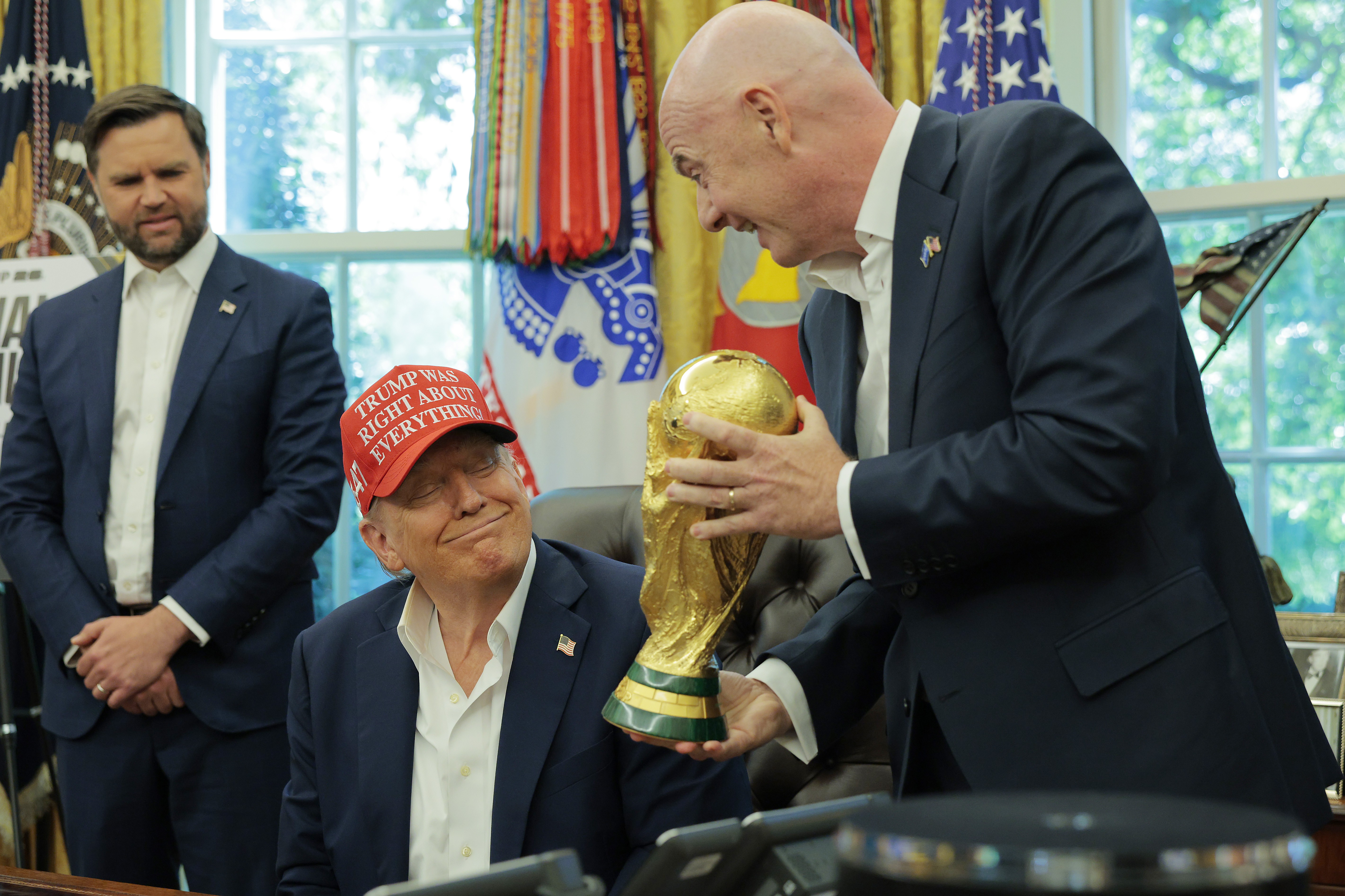 Donald Trump, de boné vermelho Trump Was Right About Everything, sorri enquanto Gianni Infantino, presidente da FIFA, lhe entrega a taça da Copa do Mundo. Um homem de terno azul observa ao fundo, em um escritório com janelas e bandeiras