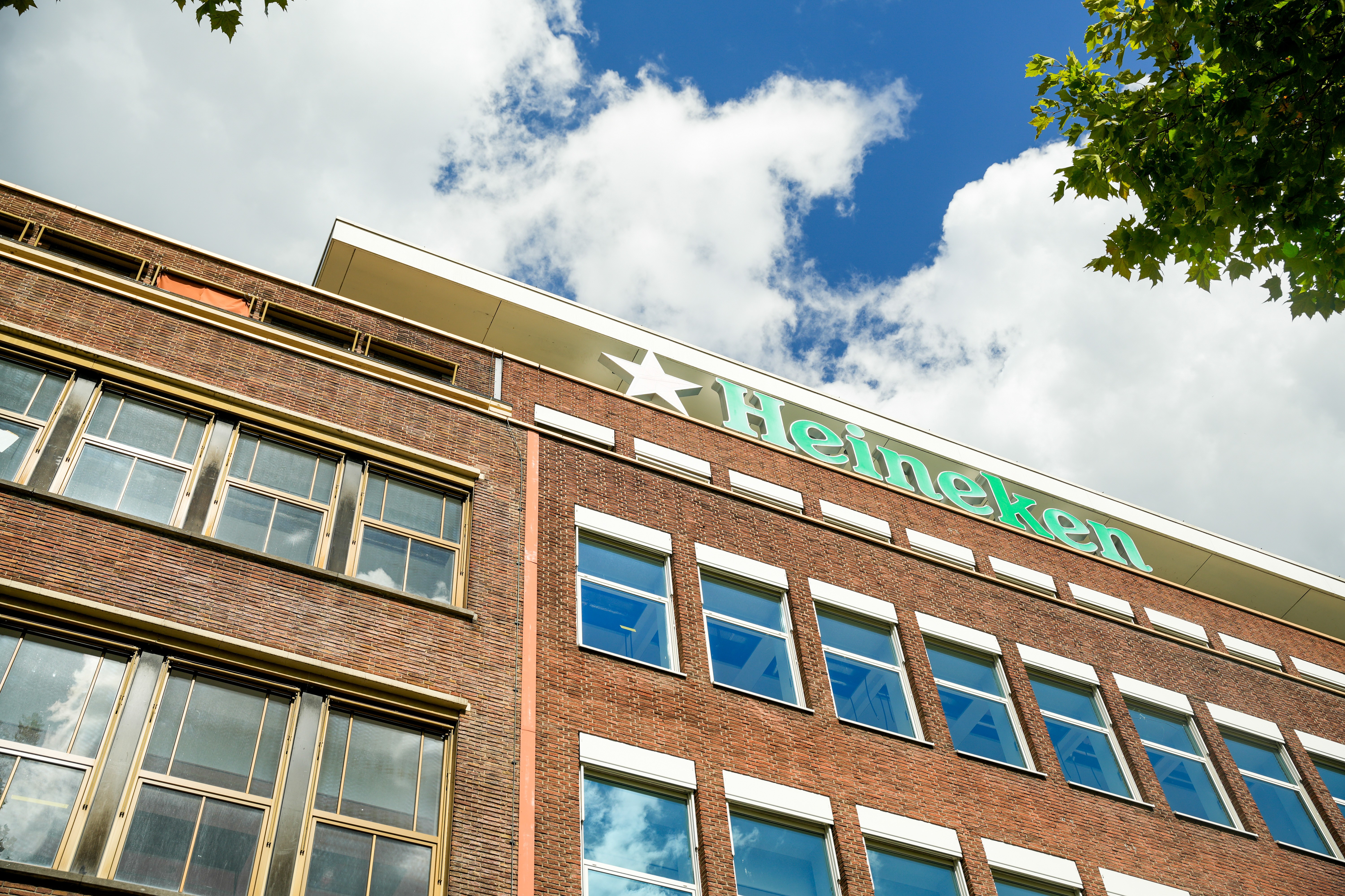 Fachada de um prédio de tijolos marrons com o logotipo verde da Heineken e uma estrela branca no topo, sob um céu azul com nuvens e folhas de árvore no canto superior direito