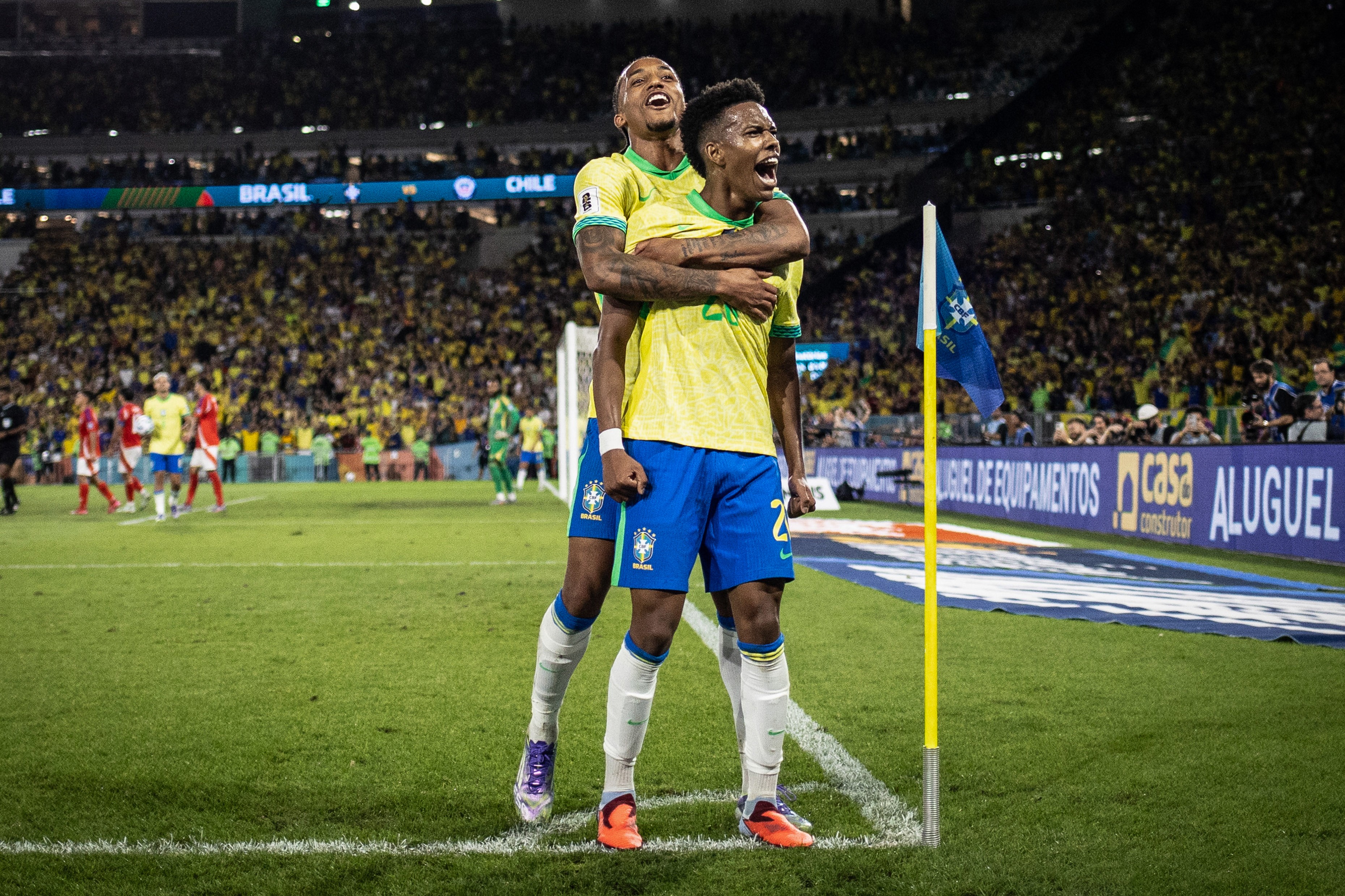 Dois jogadores de futebol brasileiros, um abraçando o outro por trás, comemoram um gol em campo, ambos sorrindo e gritando. Usam camisas amarelas e shorts azuis, em um estádio lotado de torcedores. Uma bandeira azul está à direita