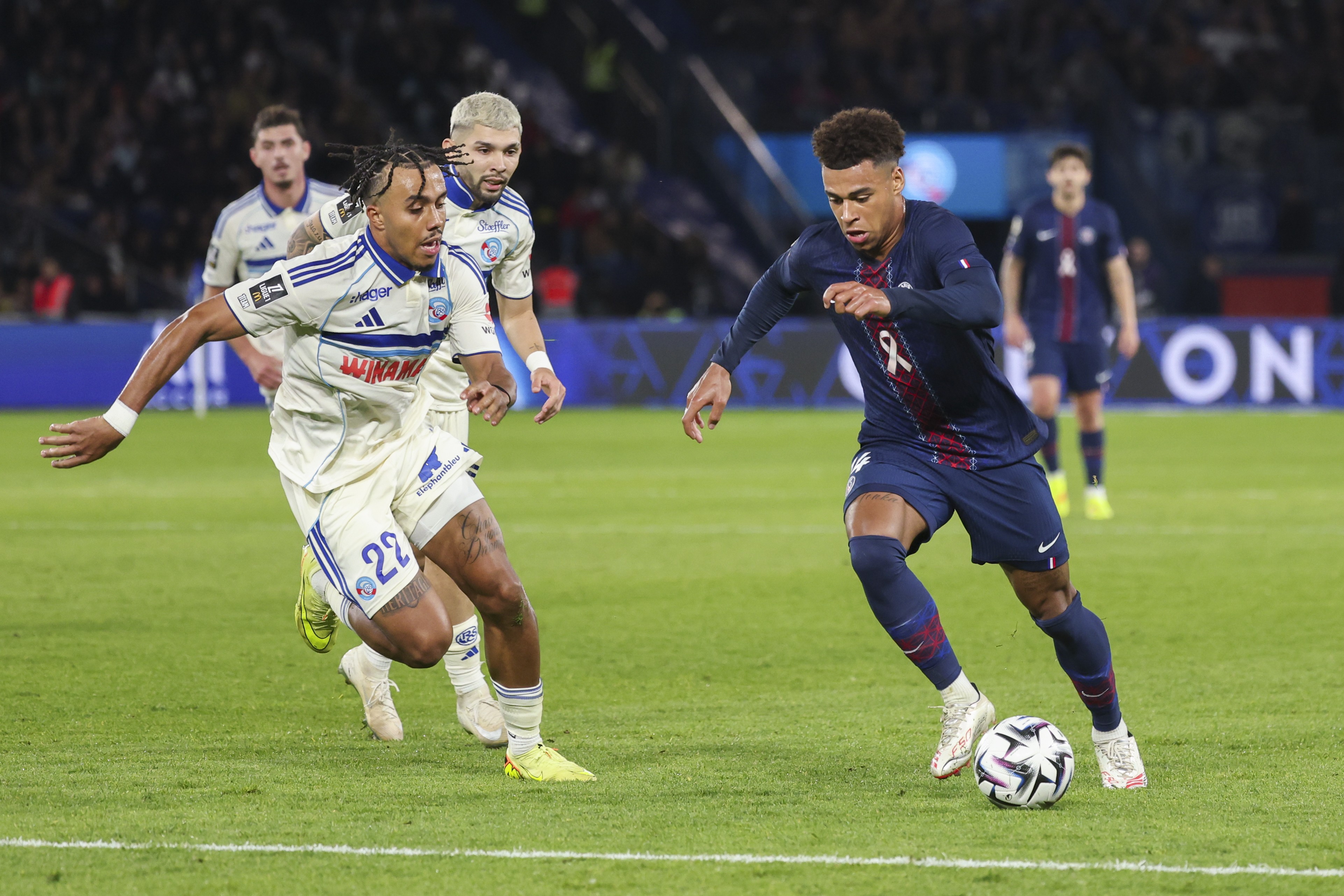 Dois jogadores de futebol correm no campo verde, um de camisa branca com listras azuis e o outro de camisa azul-marinho com faixa vermelha, driblando a bola. Outros jogadores estão ao fundo, em um estádio iluminado à noite