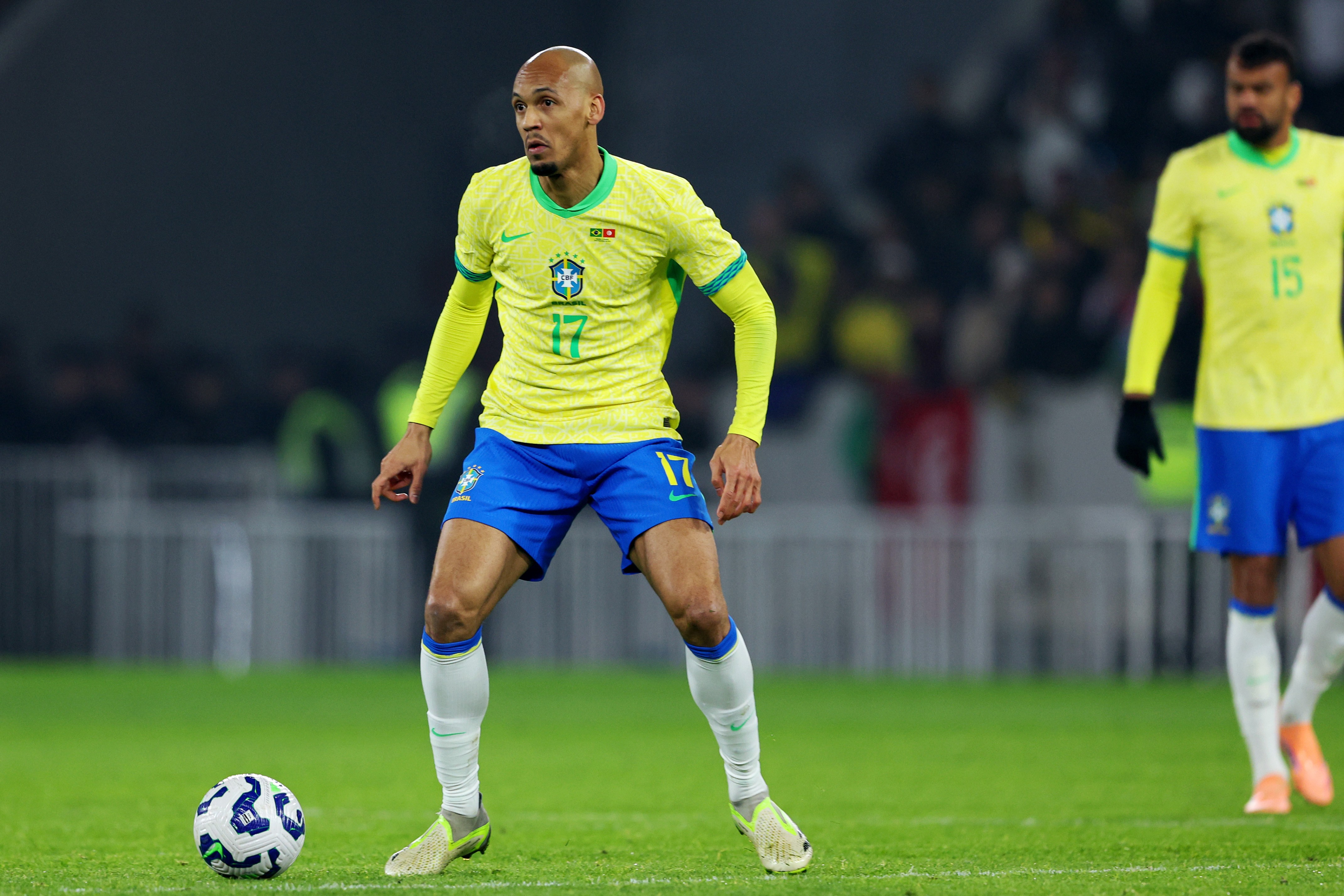 Jogador de futebol Fabinho, careca, vestindo camisa amarela de manga longa e shorts azuis com o número 17, parado em campo verde com uma bola branca e azul aos seus pés. Outro jogador desfocado aparece ao fundo.