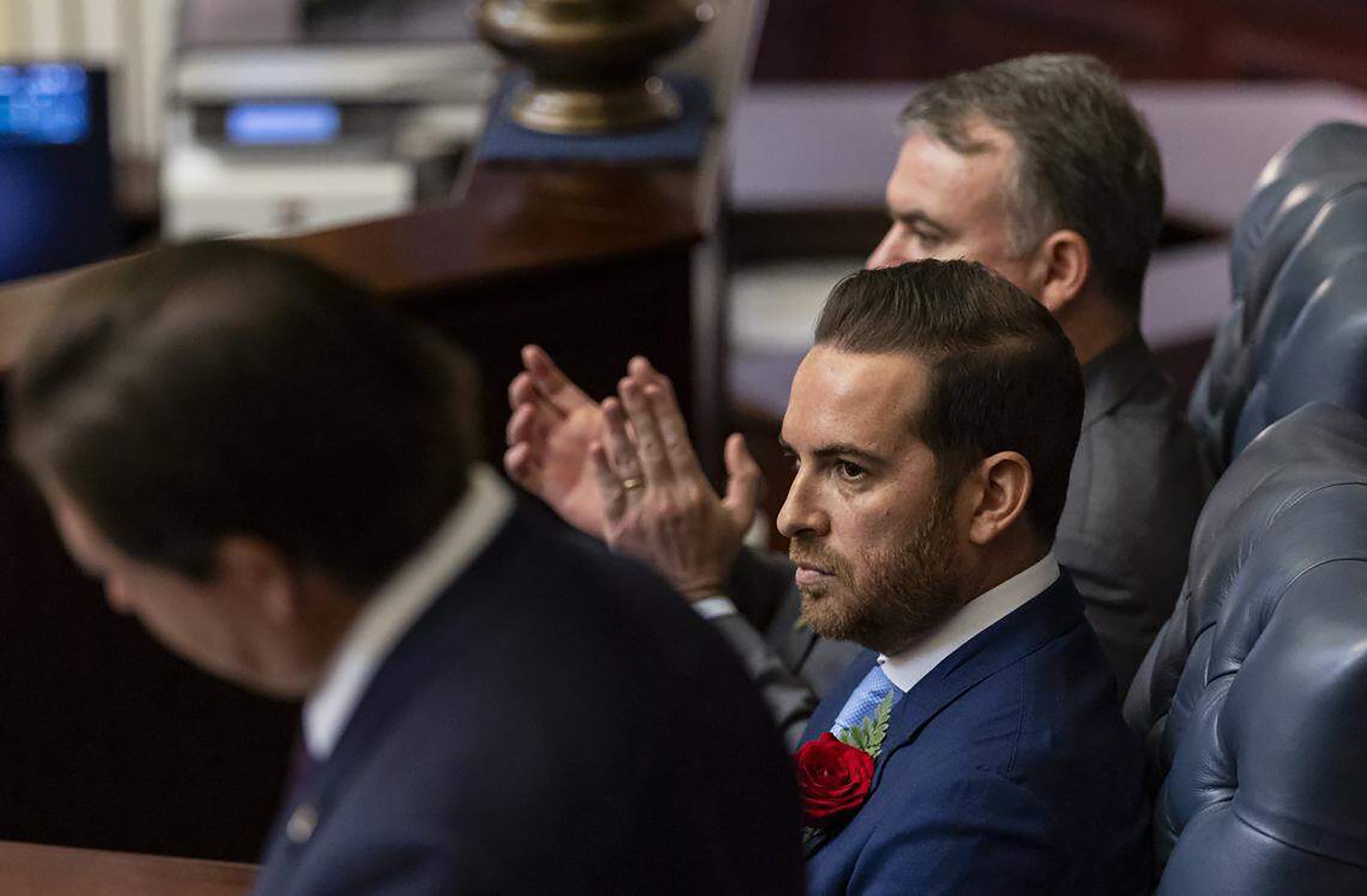 Homem barbudo de terno azul com rosa vermelha na lapela, olhando sério para a direita, enquanto outro homem atrás aplaude. Um terceiro homem, de costas, está à esquerda.