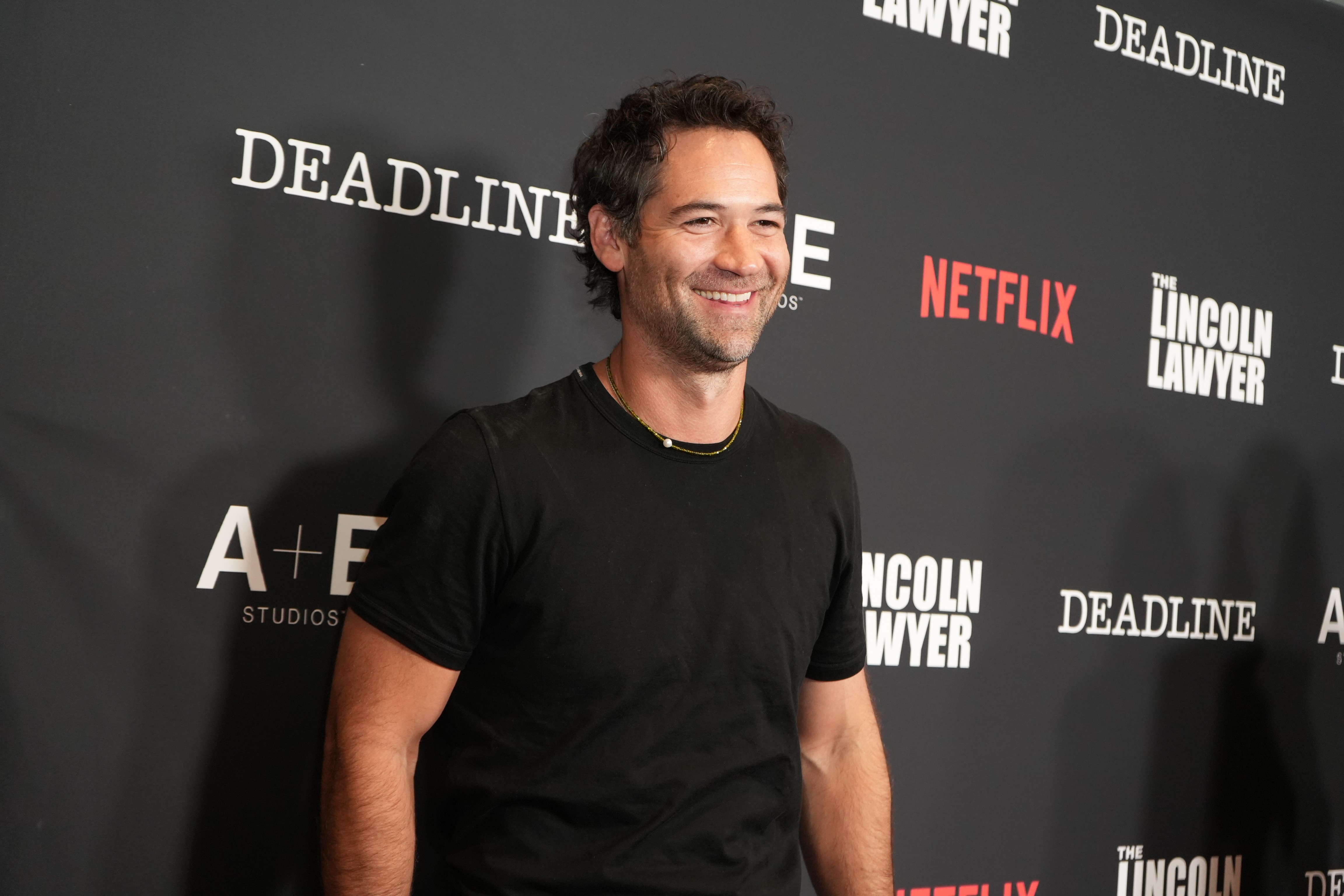 O ator Manuel Garcia-Rulfo sorri, usando uma camiseta preta e um colar dourado, em frente a um painel preto com logotipos brancos e vermelhos de Deadline, Netflix, The Lincoln Lawyer e A+E Studios