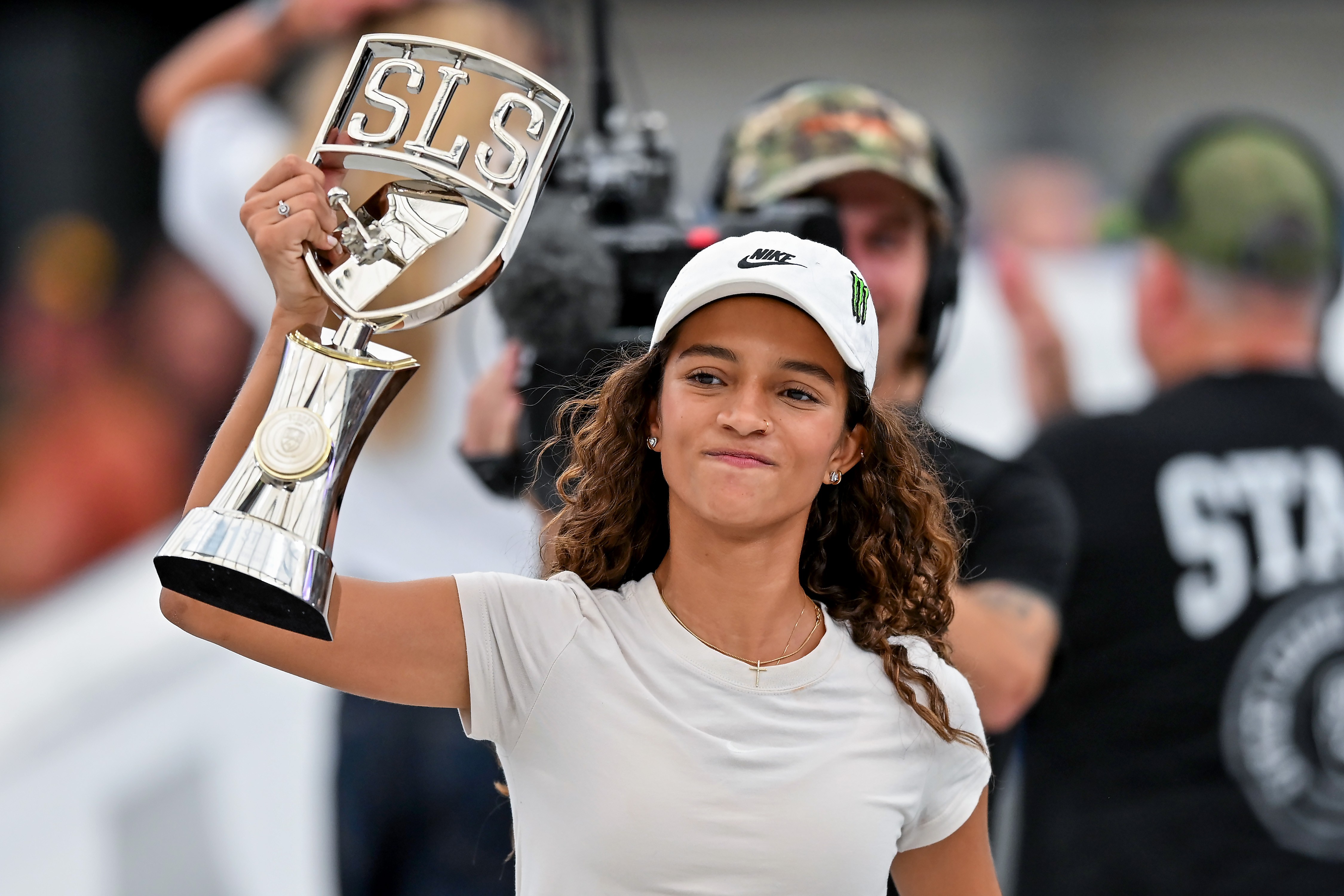 Jovem skatista Rayssa Leal, com cabelos cacheados e boné branco, sorri segurando um troféu prateado com a sigla SLS em relevo, celebrando uma vitória