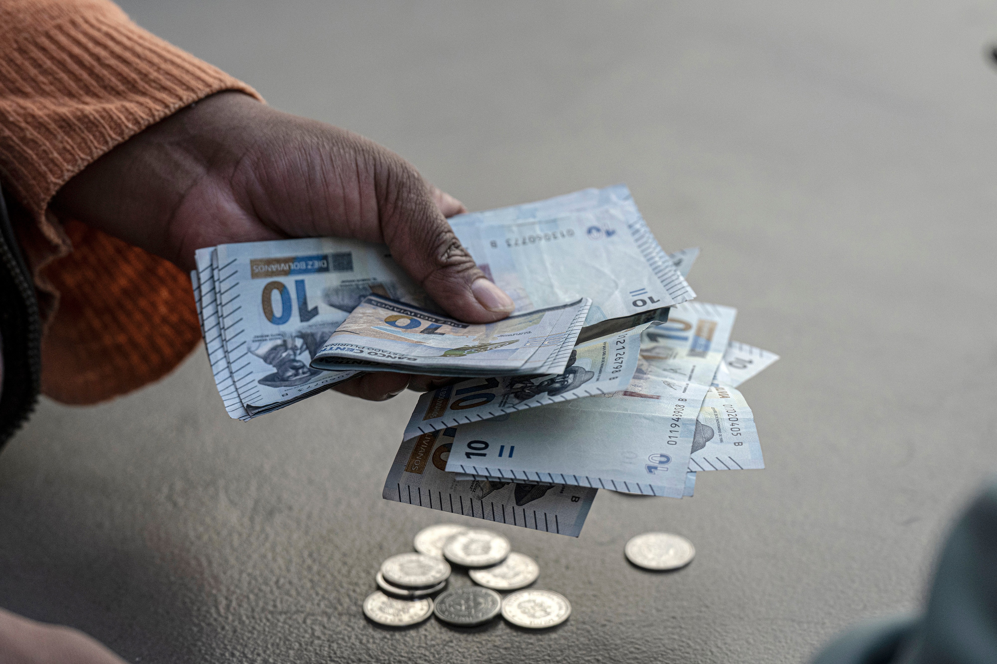 Mão de pele escura segurando várias notas de 10, com moedas espalhadas sobre uma superfície cinza