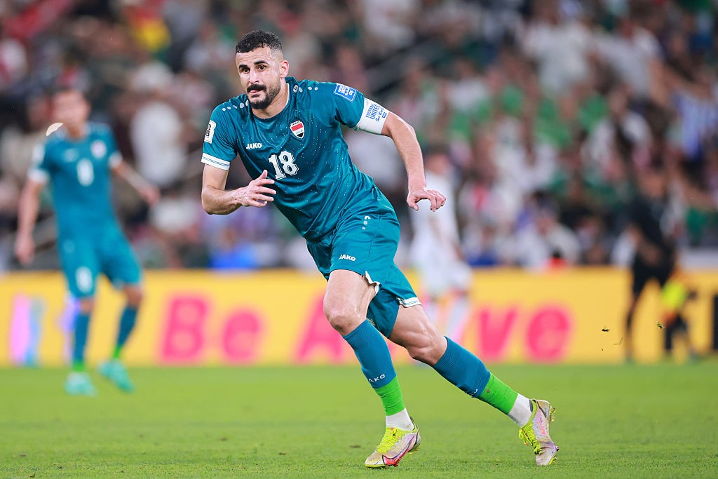 Jogador de futebol com barba e bigode, vestindo uniforme verde-azulado com o número 18, correndo no campo gramado de um estádio lotado