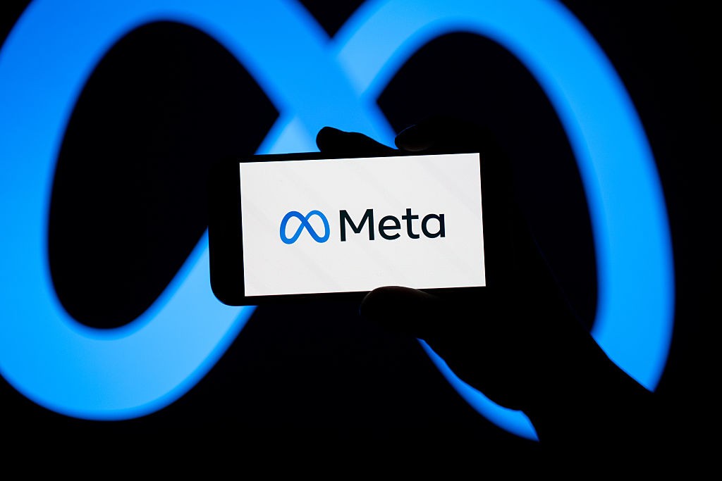 Mão segurando um celular com o logo da Meta em azul e branco, sobre um fundo escuro com o símbolo do infinito em neon azul