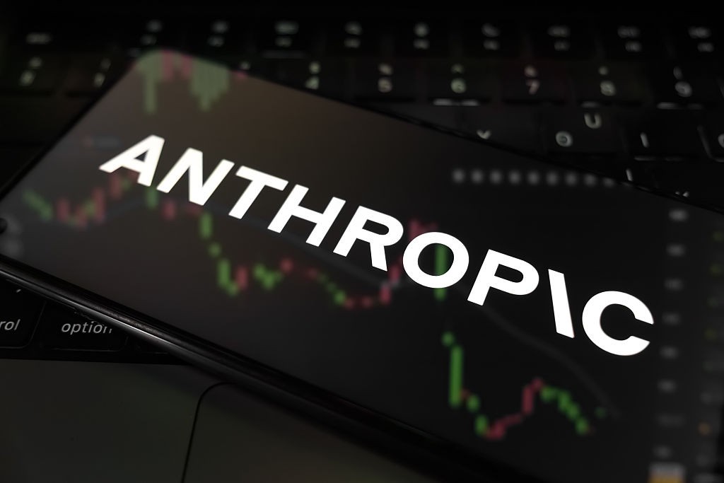 A palavra "ANTHROPIC" em letras brancas maiúsculas aparece na tela de um smartphone escuro, com gráficos de linhas verdes e vermelhas no fundo, sobre um teclado de notebook preto