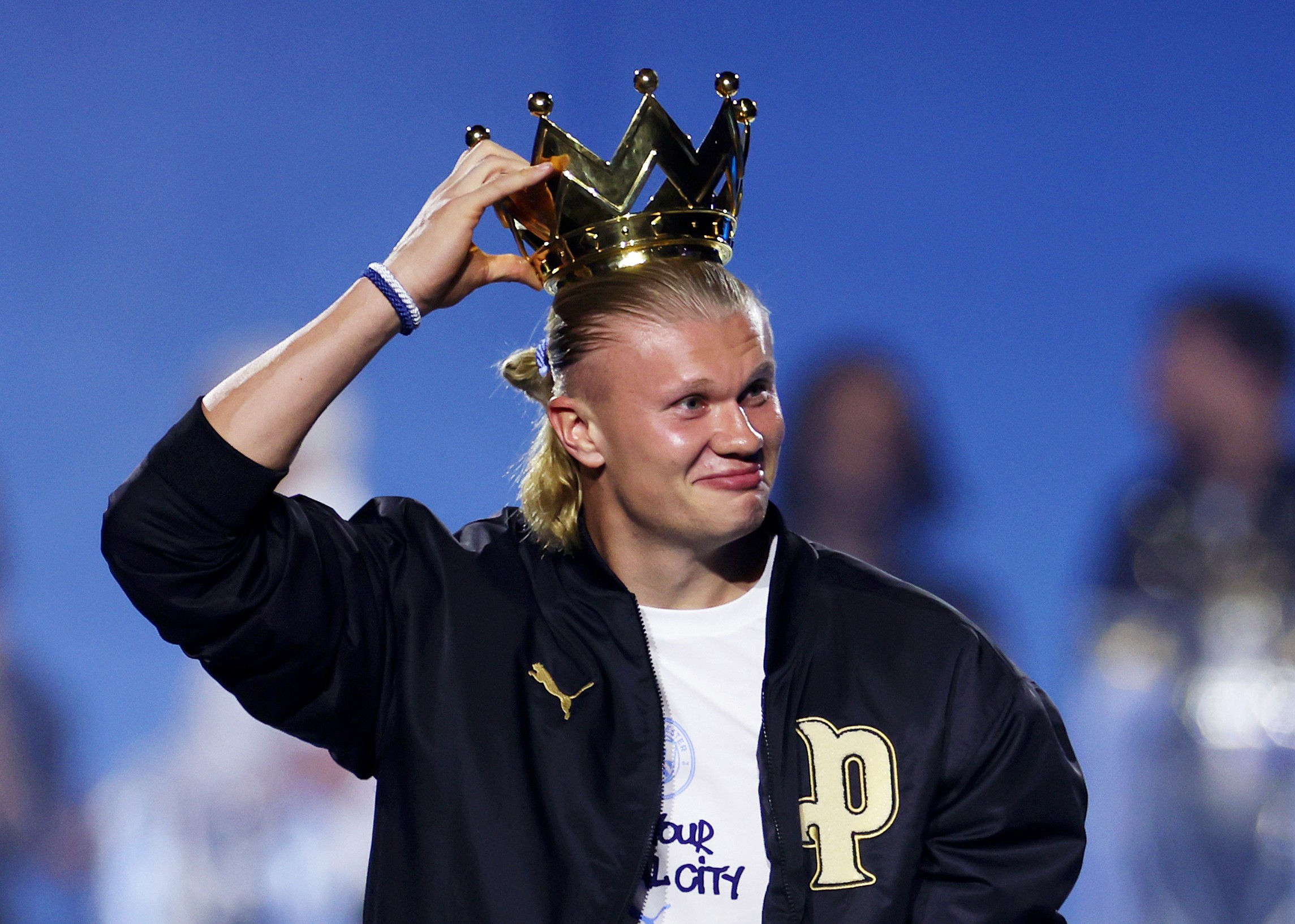 Jogador Erling Haaland, loiro de cabelo preso, sorri enquanto ajusta uma coroa dourada na cabeça, vestindo jaqueta preta com logo Puma e camiseta branca, em fundo azul desfocado