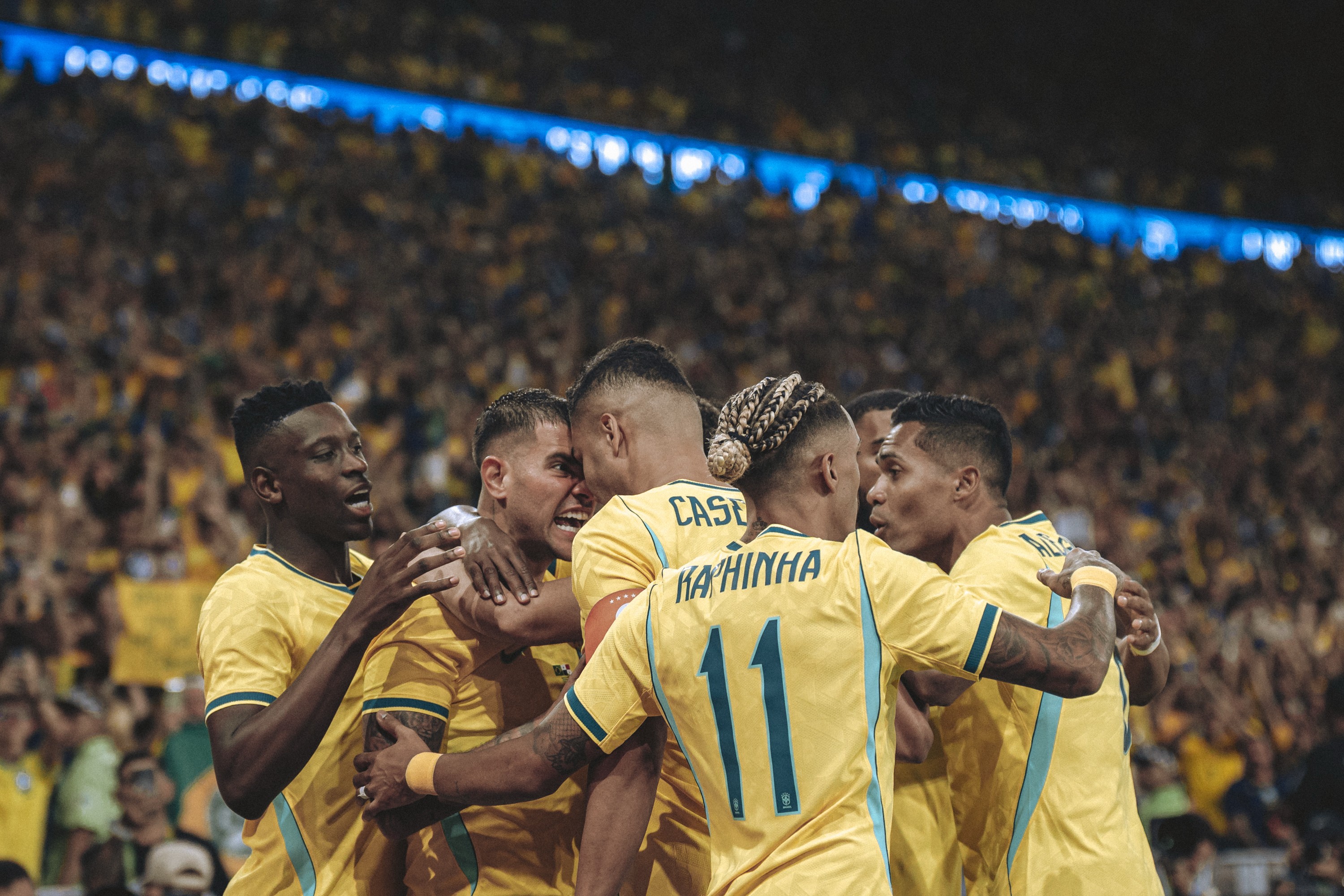 Jogadores de futebol brasileiros em camisas amarelas e azuis se abraçam em comemoração no campo, com a torcida ao fundo