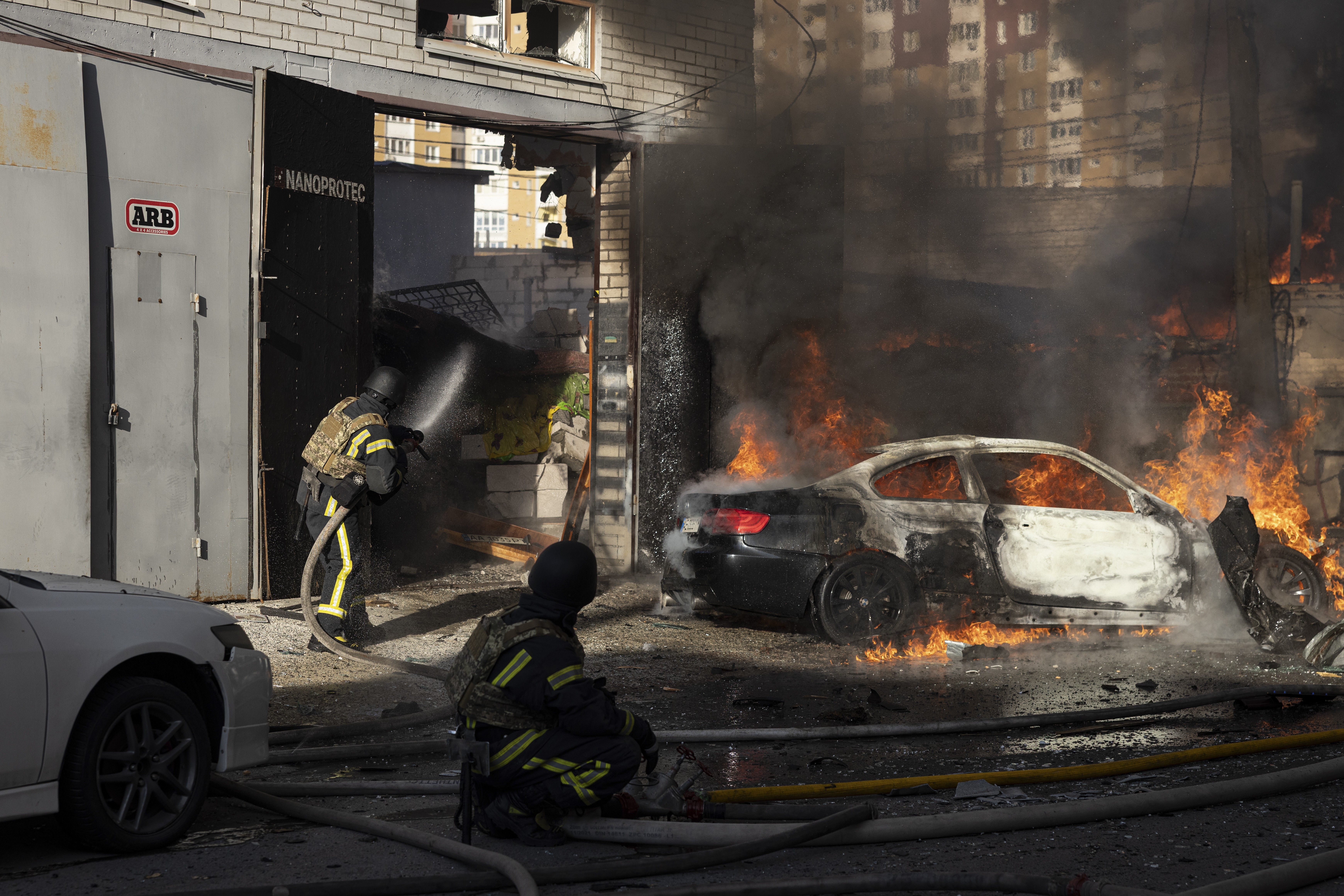Dois bombeiros em uniformes escuros com faixas amarelas combatem um incêndio em um carro preto parcialmente destruído, com chamas e fumaça densa, em frente a um prédio danificado em Kiev. Um bombeiro está de pé, usando uma mangueira, enquanto outro está agachado no chão, observando. Um carro branco está estacionado à esquerda.
