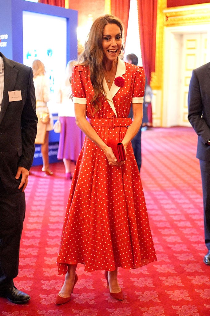 Kate Middleton sorrindo, usando um vestido midi vermelho de bolinhas brancas, gola e punhos brancos, cinto e broche de flor vermelha, segurando uma clutch vermelha, em um salão com carpete vermelho