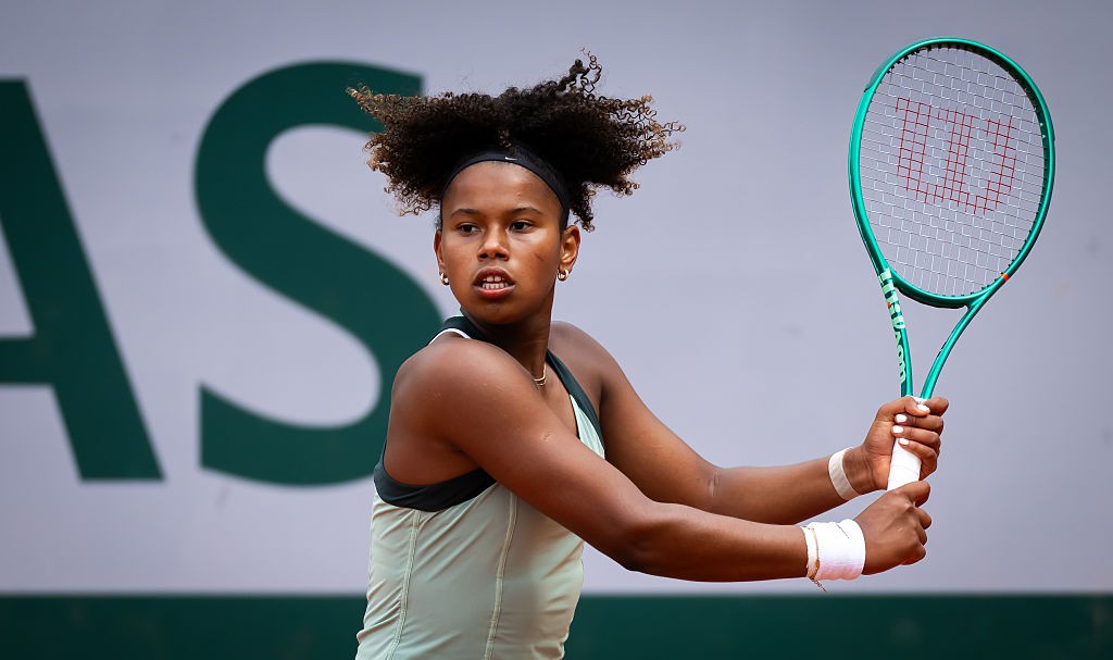 Jovem tenista negra com cabelo crespo preso por uma faixa preta, vestindo regata verde-clara, segurando uma raquete de tênis verde-água com as duas mãos e munhequeiras brancas, em quadra de saibro com fundo branco e letras verdes
