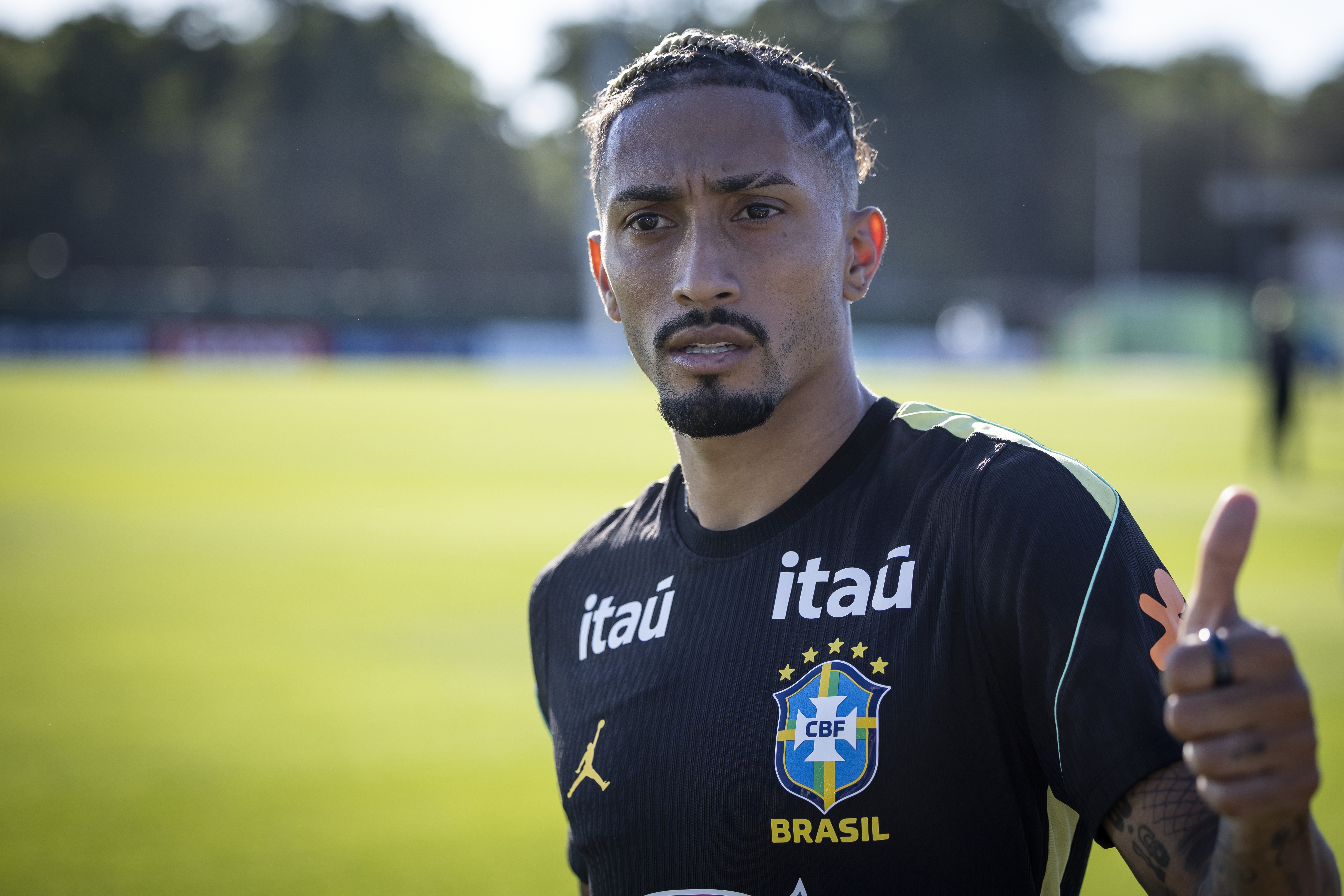 Marquinhos, jogador de futebol, em campo com camisa preta da seleção brasileira, barba e cabelo trançado, fazendo sinal de positivo com a mão direita