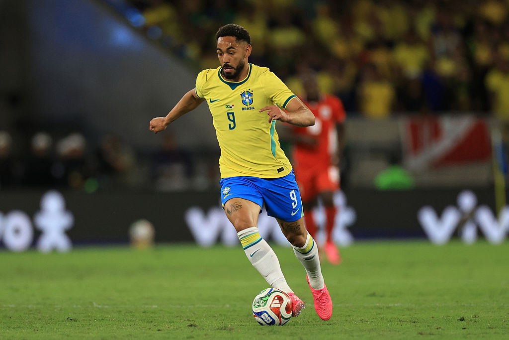 Jogador de futebol brasileiro, com barba, vestindo camisa amarela e calção azul com o número 9, driblando a bola no campo verde de um estádio, com torcedores borrados ao fundo