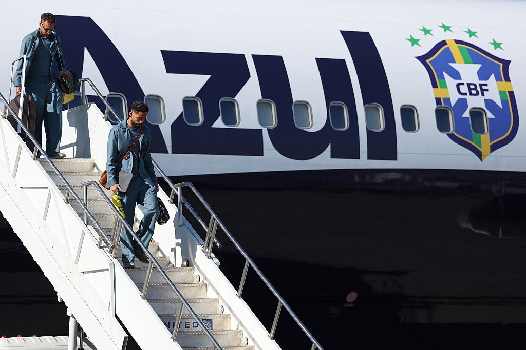 Dois homens descem a escada de um avião branco com o logotipo "AZUL" e o escudo da CBF, com cinco estrelas verdes acima
