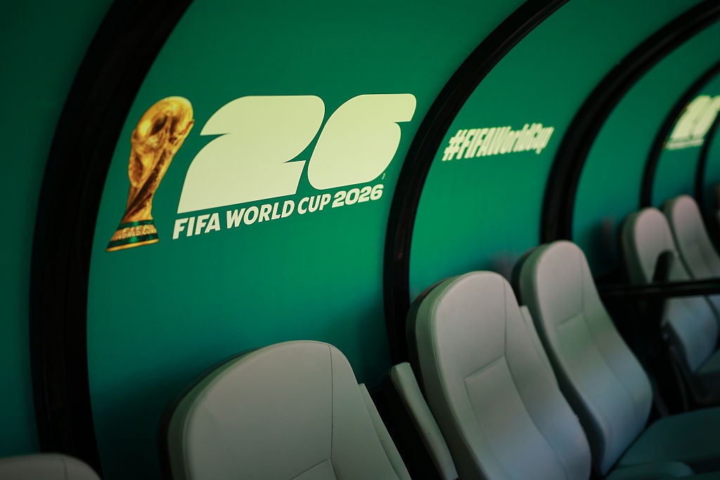 Bancos cinzas de um banco de reservas de futebol, com a parede verde-água ao fundo exibindo o troféu da Copa do Mundo e o texto "26 FIFA WORLD CUP 2026" e "#FIFAWorldCup"