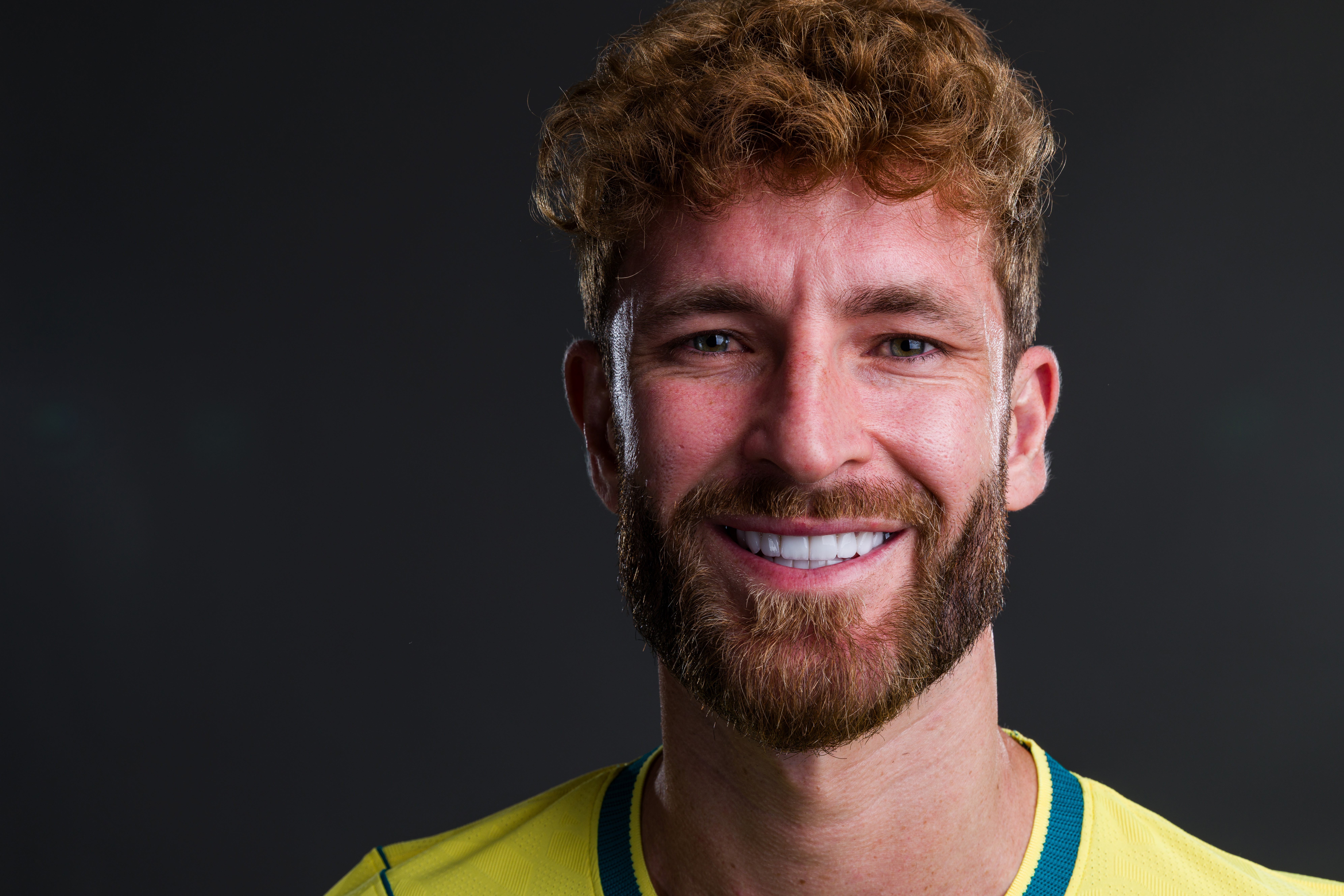 Homem branco de cabelo e barba ruivos, encaracolados, sorrindo com dentes brancos e alinhados, vestindo camisa amarela com detalhes azuis, em fundo cinza escuro