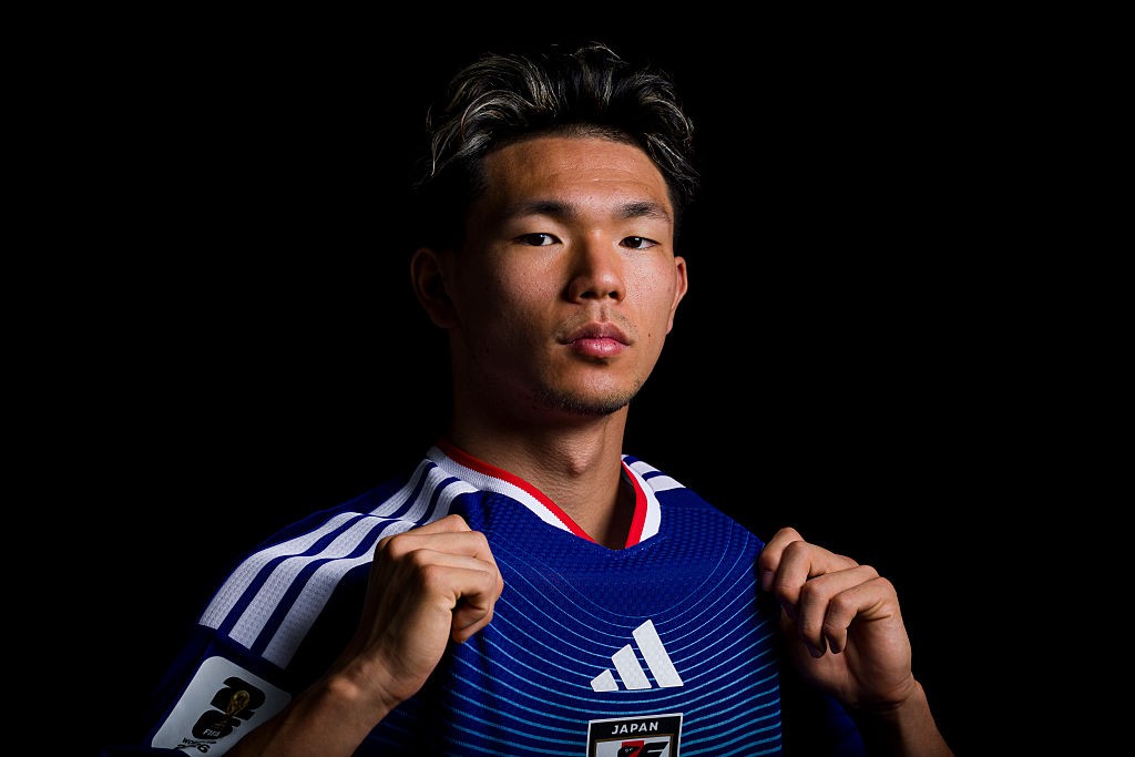 Jovem jogador de futebol japonês, com cabelo escuro e mechas claras, vestindo uma camisa azul da seleção do Japão com listras brancas nos ombros e gola vermelha, segurando a gola com as duas mãos, em fundo preto