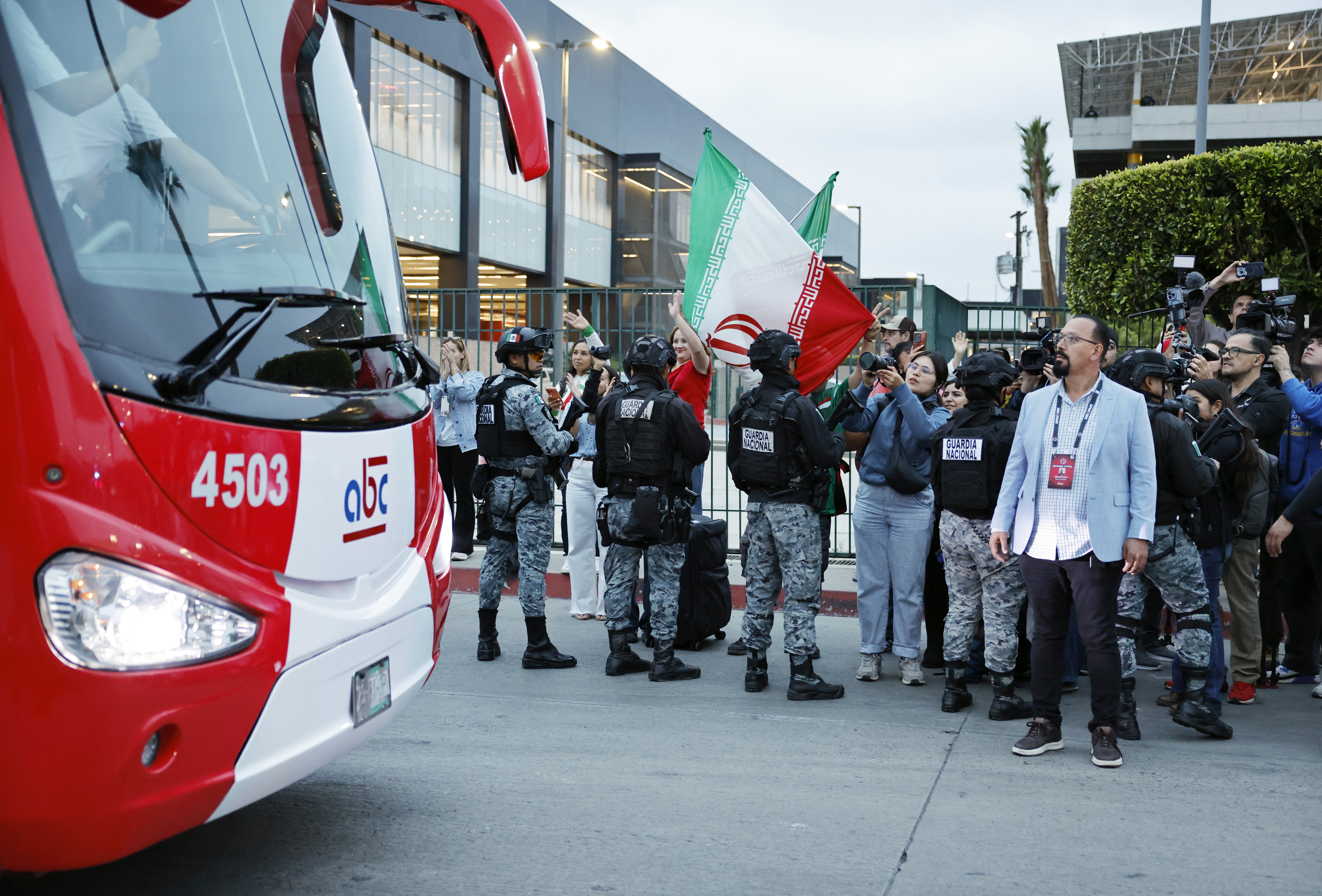 Ônibus vermelho e branco com número 4503 na lateral, e um grupo de pessoas, incluindo membros da Guarda Nacional em uniforme camuflado, e civis, alguns segurando a bandeira do Irã, em frente a um prédio moderno. Um homem de óculos e blazer azul claro se destaca no centro, olhando para cima. O céu está nublado