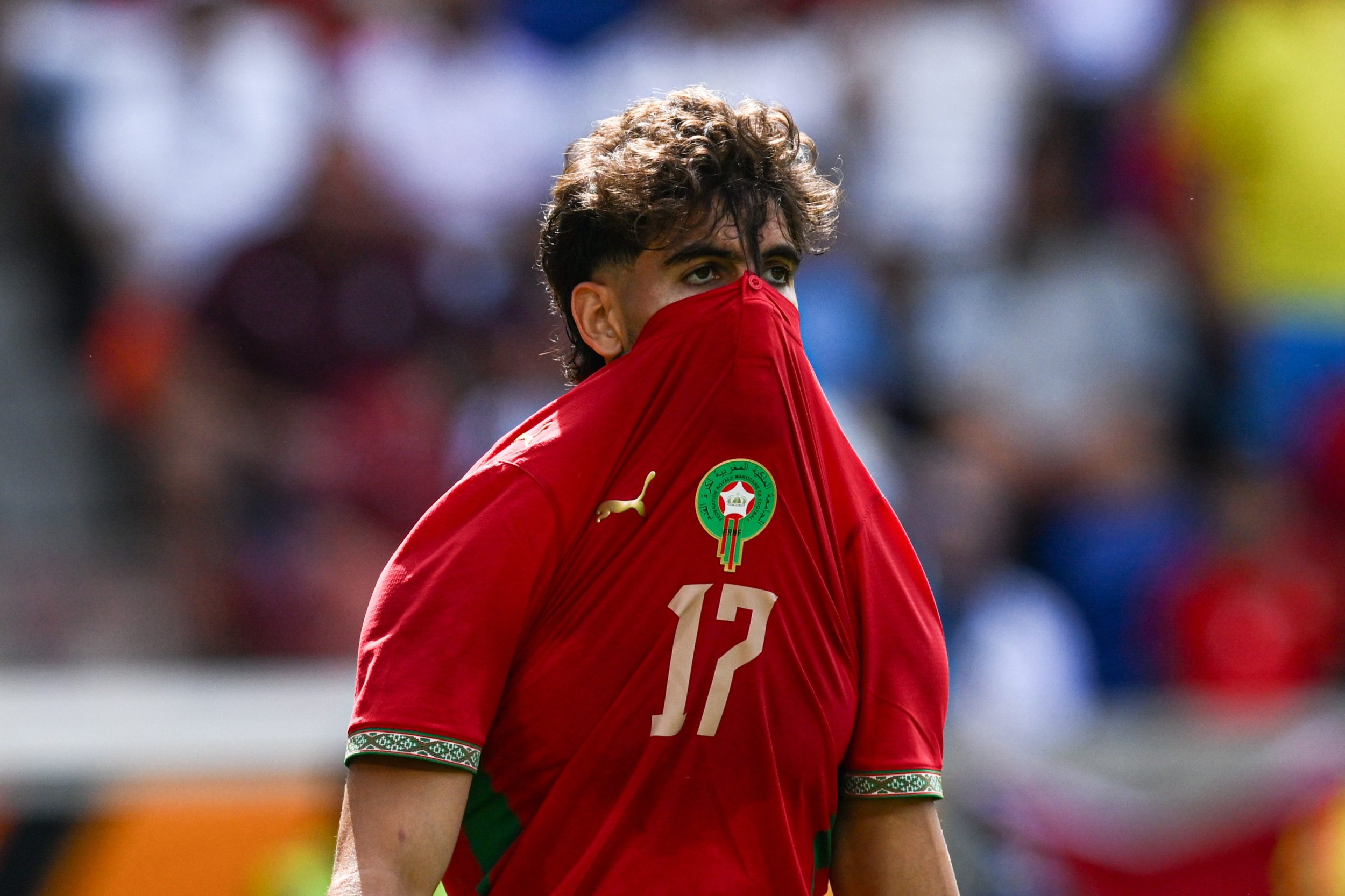 Jovem jogador de futebol com cabelo castanho encaracolado, vestindo camisa vermelha com o número 17 e o emblema de Marrocos, cobrindo o rosto com a gola da camisa, em um campo de futebol